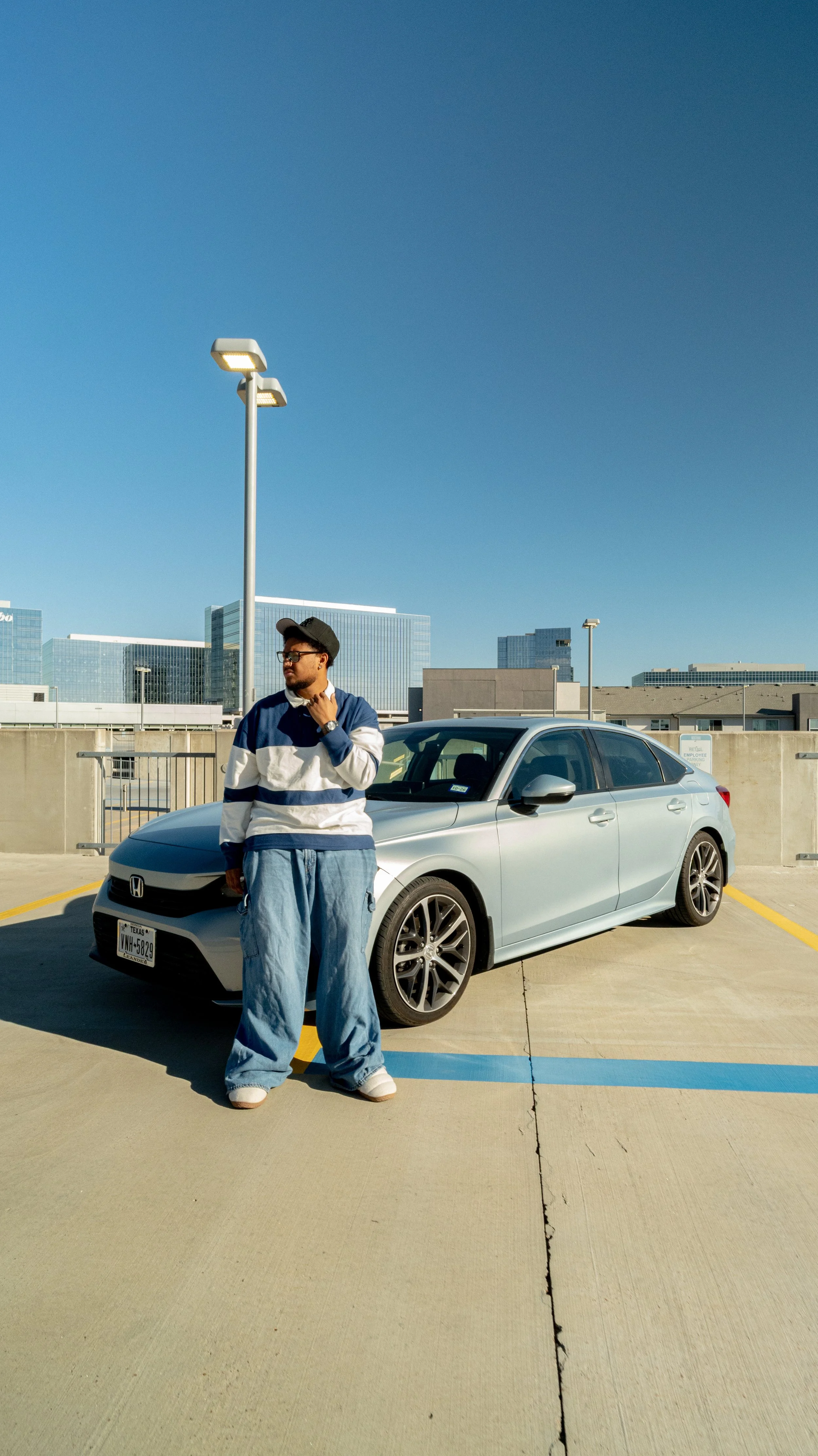 A man in oversized jeans, a striped jacket, and a cap stands next to a silver Honda sedan in a parking lot with a city skyline in the background.
