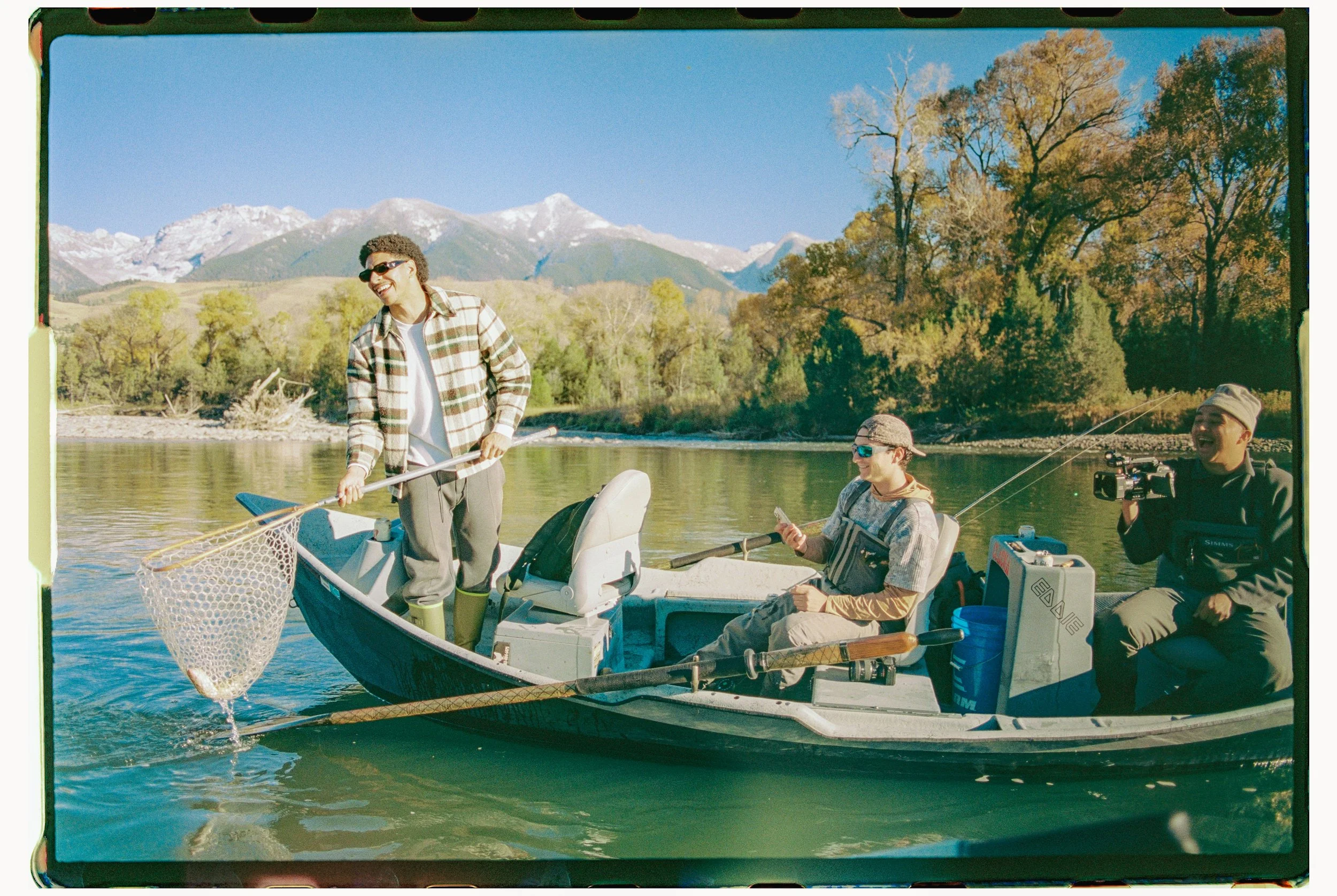 Three men on a boat fishing on a river with mountains and trees in the background, one man is standing with a fishing net, another man is sitting holding a phone, and the third man is sitting holding a camera.