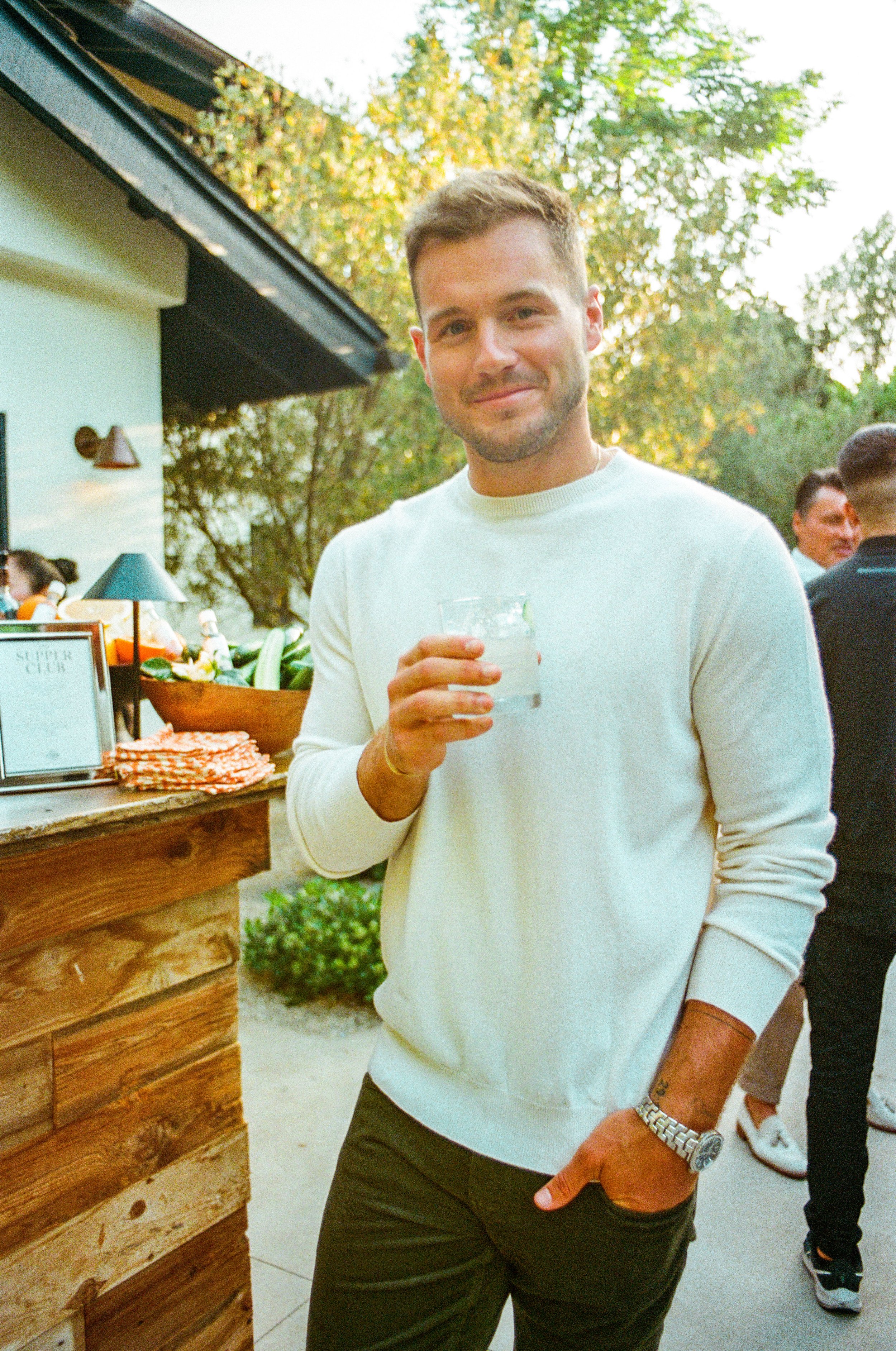 A man outdoors at a social event holding a glass of drink, wearing a white sweater and a watch, standing next to a wooden bar with a sign and snacks.