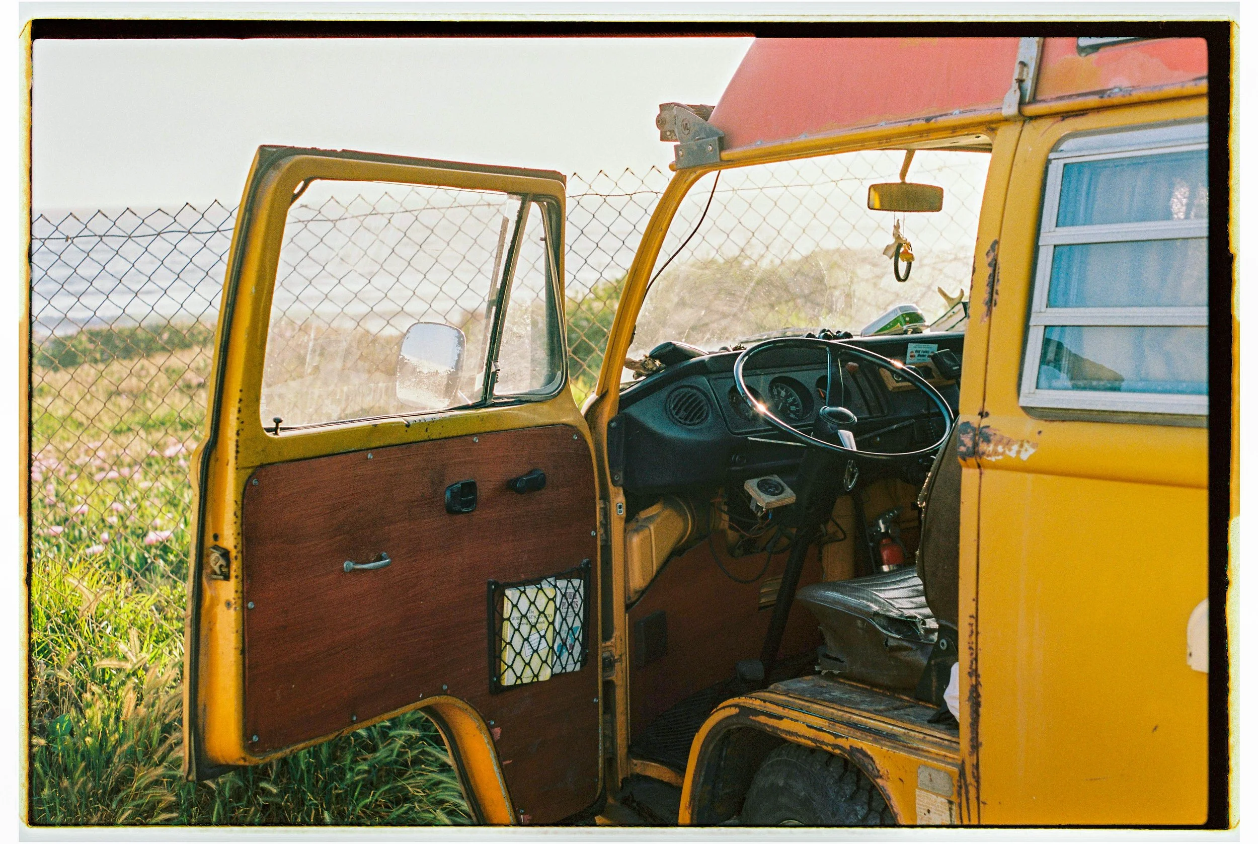 Interior of a yellow vintage vehicle with a wooden panel on the door, featuring an open door, a steering wheel, dashboard, and side window, with a field and fence in the background.