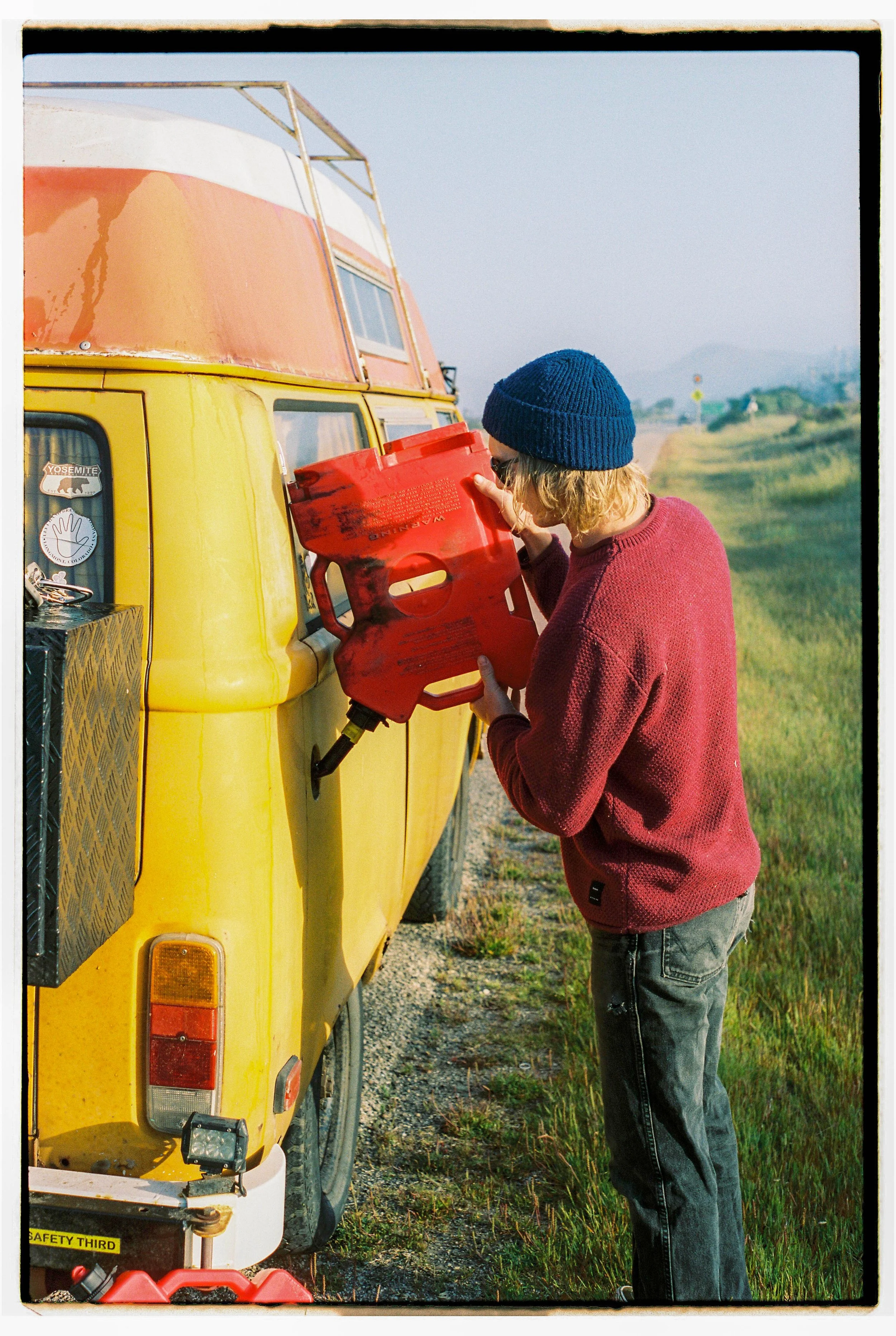 A young man with blond hair, wearing a blue beanie, sunglasses, a red sweater, and jeans, refueling a yellow van with a red gas canister on a rural roadside with green fields and distant mountains.