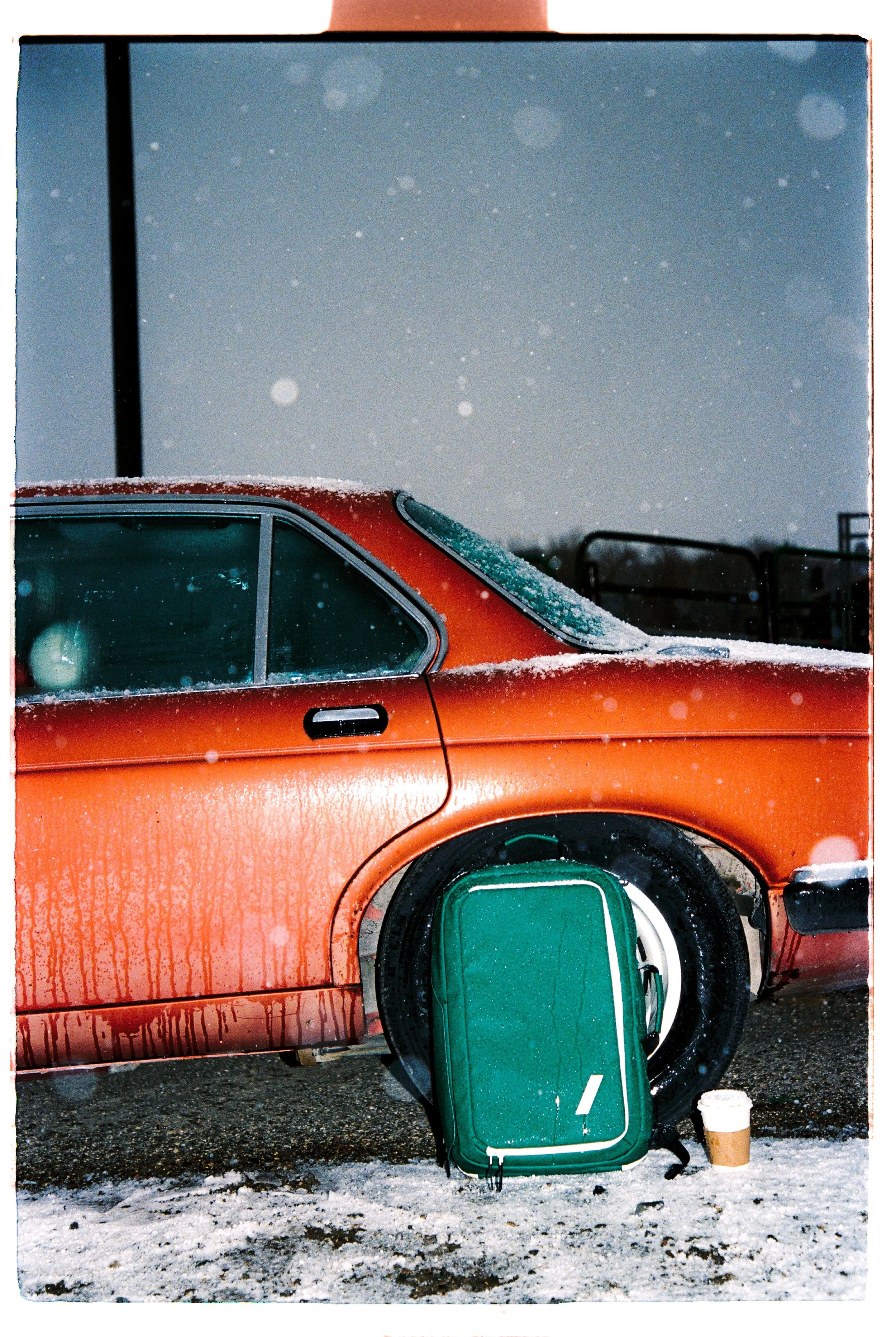 An orange car covered in snow with a green suitcase leaning against the tire and a disposable coffee cup on the ground during snowy weather.