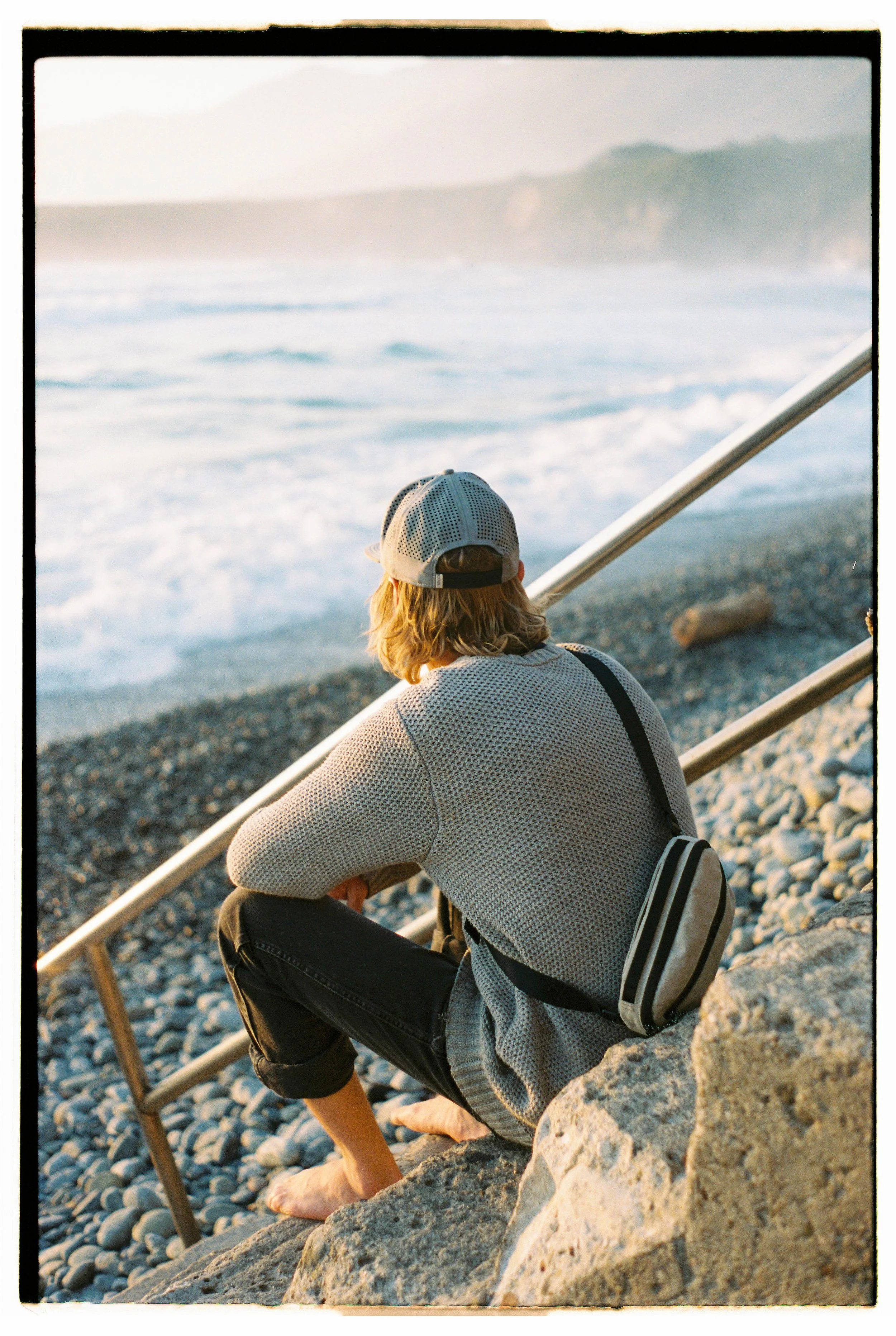 Person with blonde hair, wearing a mesh cap, long-sleeve shirt, and black pants, sitting barefoot on rocks by the beach, gazing at the ocean with waves and distant cliffs, during sunset.