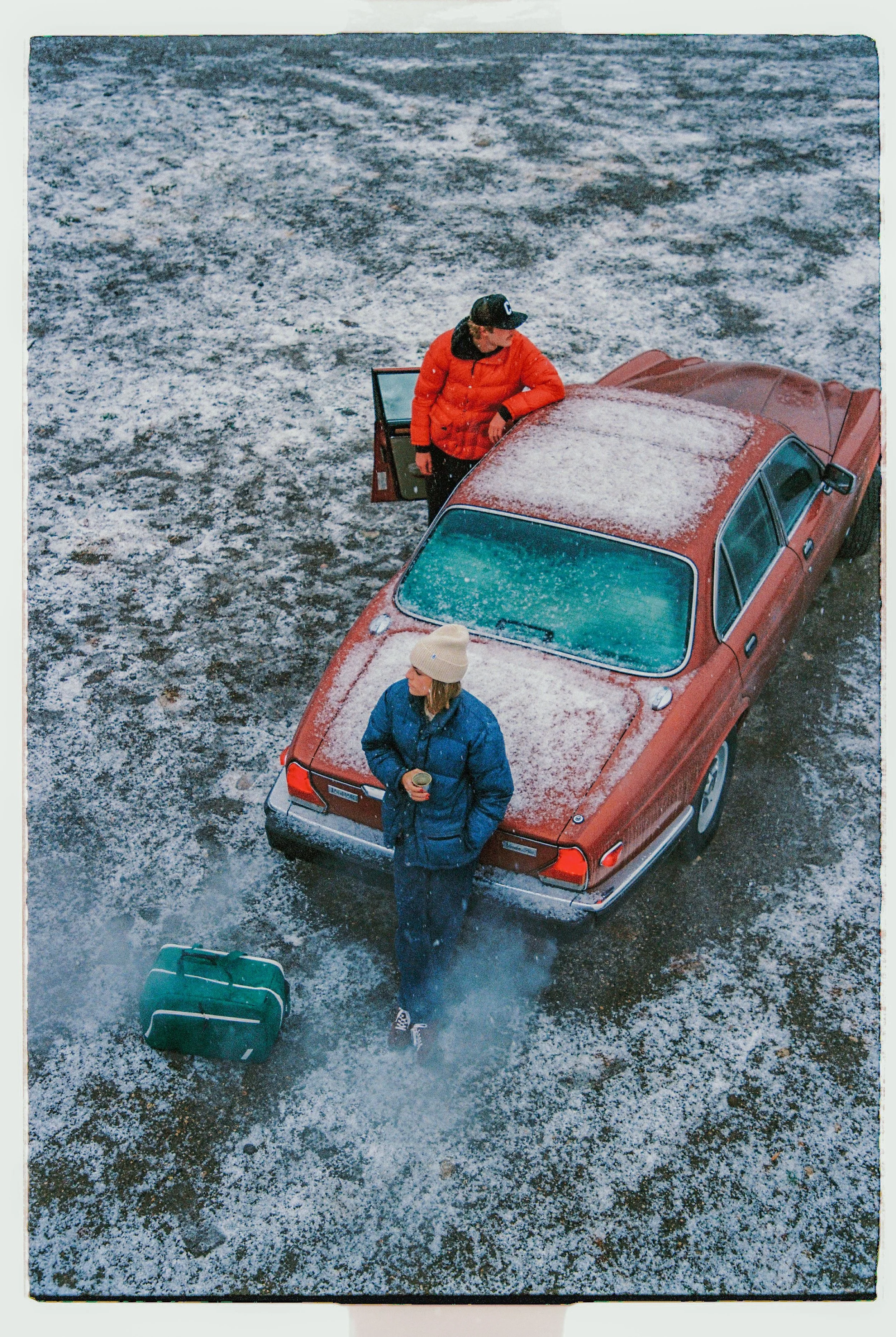 A woman in a blue winter coat and beige knit hat stands outside a red car with snow on the roof, holding a cup, while a man in an orange jacket and black cap stands with the car door open on a snow-covered ground. There is a green suitcase near their