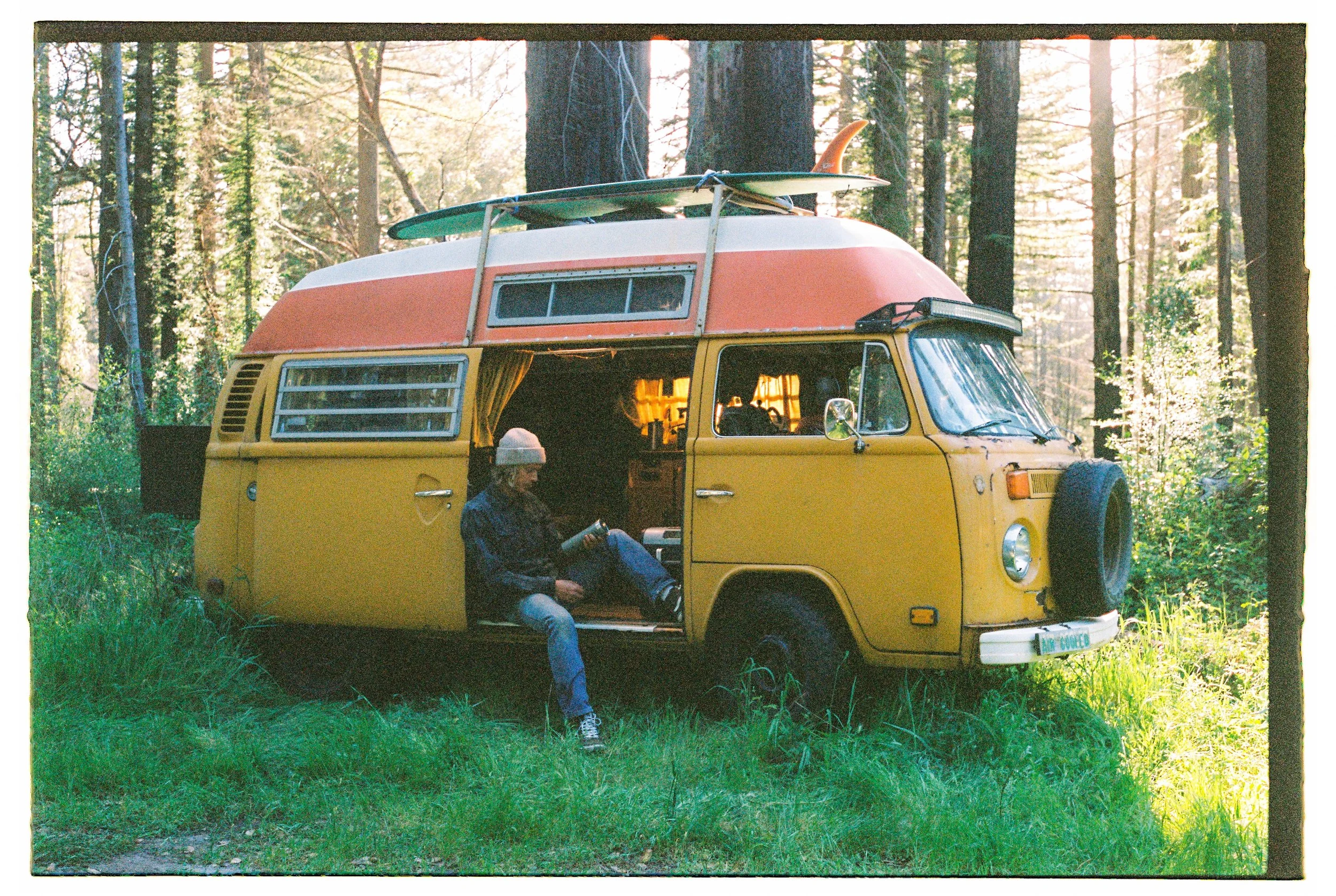 A yellow camper van with a pink and white rooftop, parked in a wooded forest. The side door is open, and a person wearing a gray beanie and jacket sits on the step outside, reading a book. Two surfboards are secured on top of the camper.
