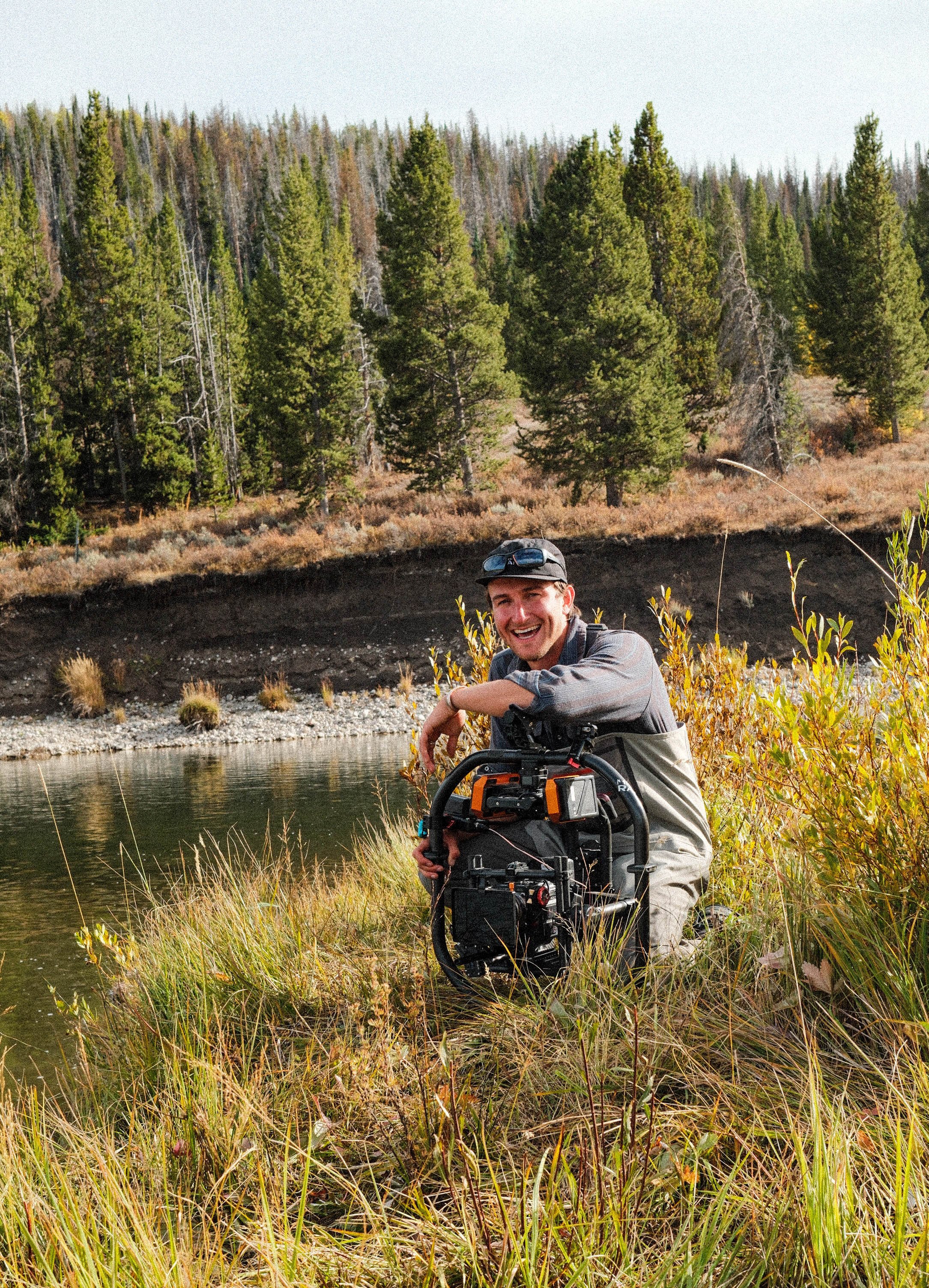 A man kneeling on the grass near a body of water, holding a professional camera rig, surrounded by trees and shrubs in a natural outdoor setting.
