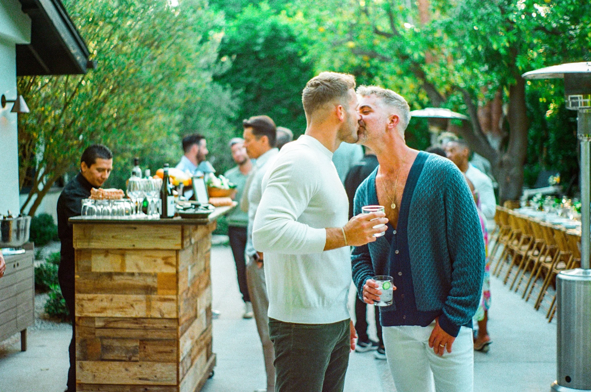 Two men are kissing each other while holding drinks at an outdoor gathering with other guests and a bar in the background.