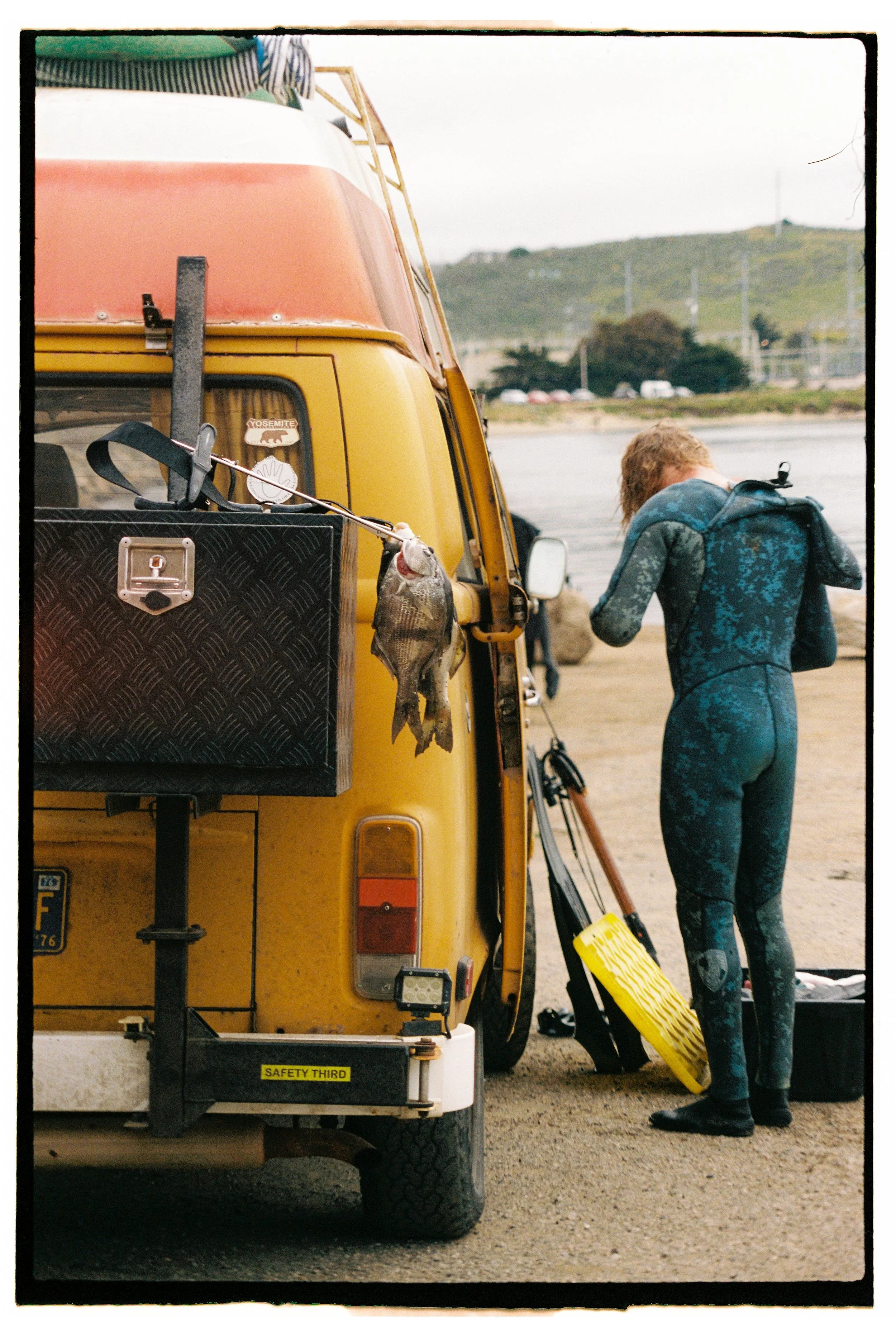 A diver in wetsuit standing next to a yellow van with a fishing rod holding a caught fish attached to its side, parked on a dirt area near a body of water with hills in the background.