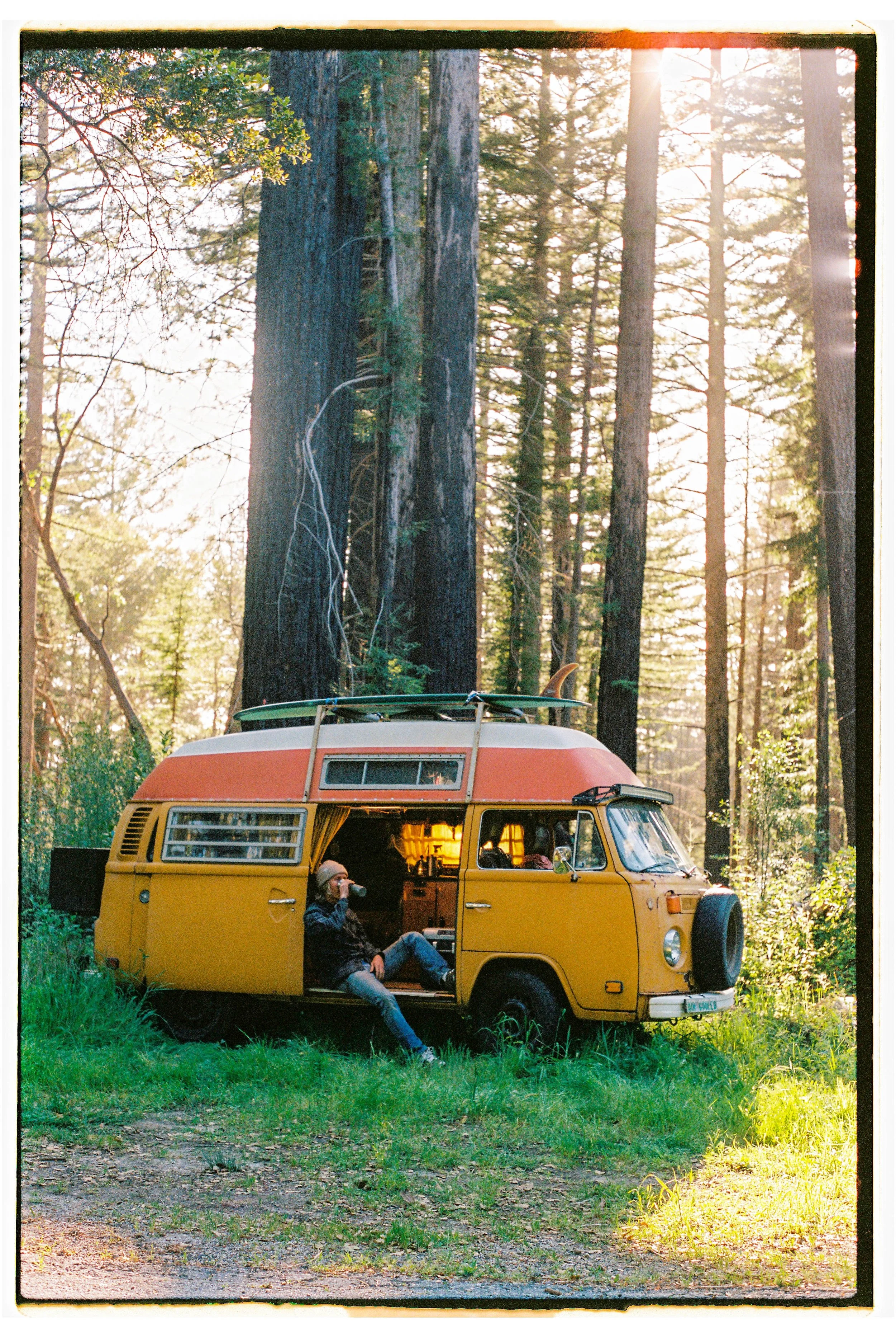 A vintage yellow and orange camper van parked in a forest clearing with tall trees. A person sitting on the van's step is drinking from a cup, surrounded by lush green grass, with sunlight filtering through the trees.