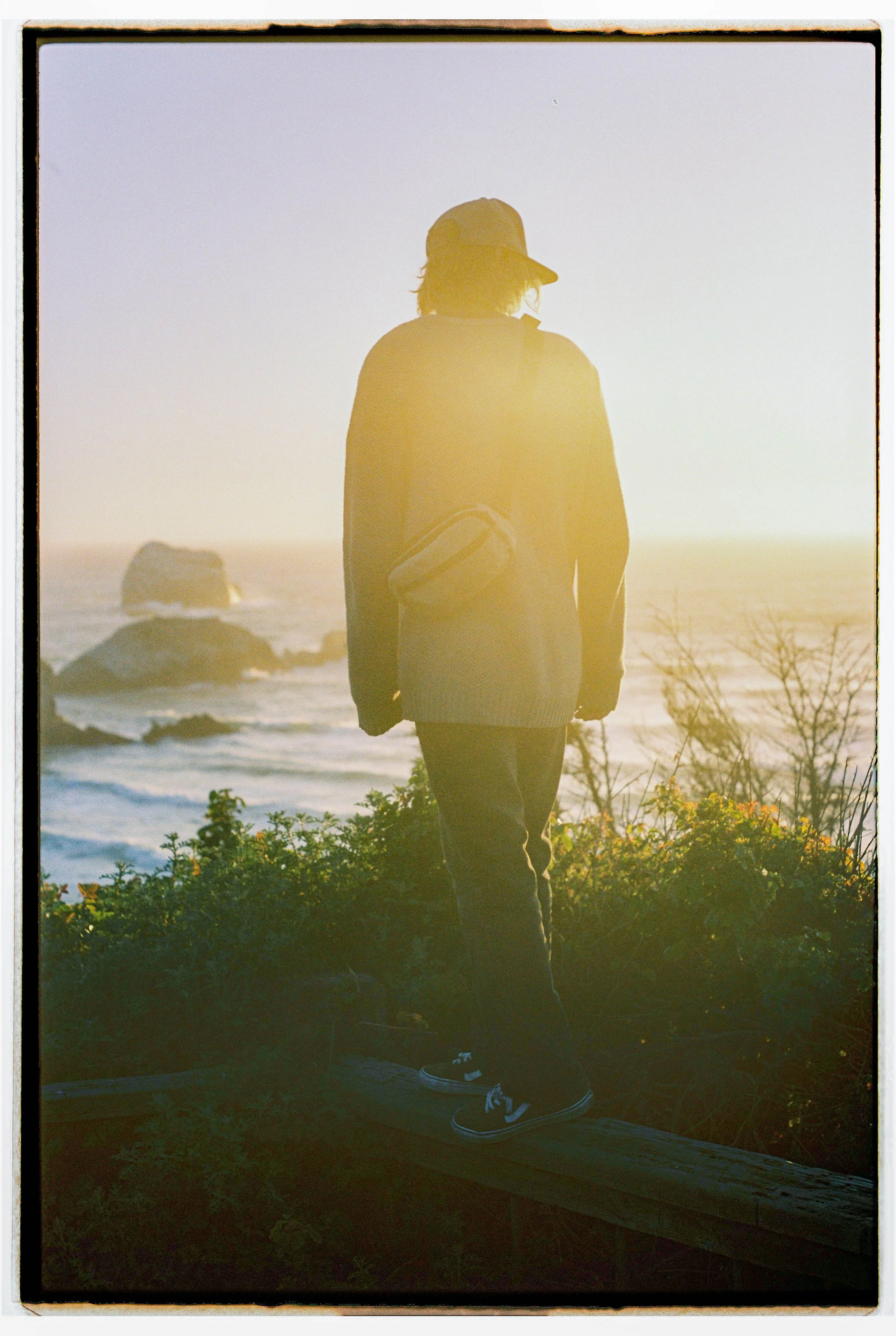 A person standing on a wooden beam overlooking the ocean at sunset, wearing a cap, jacket, and sneakers.