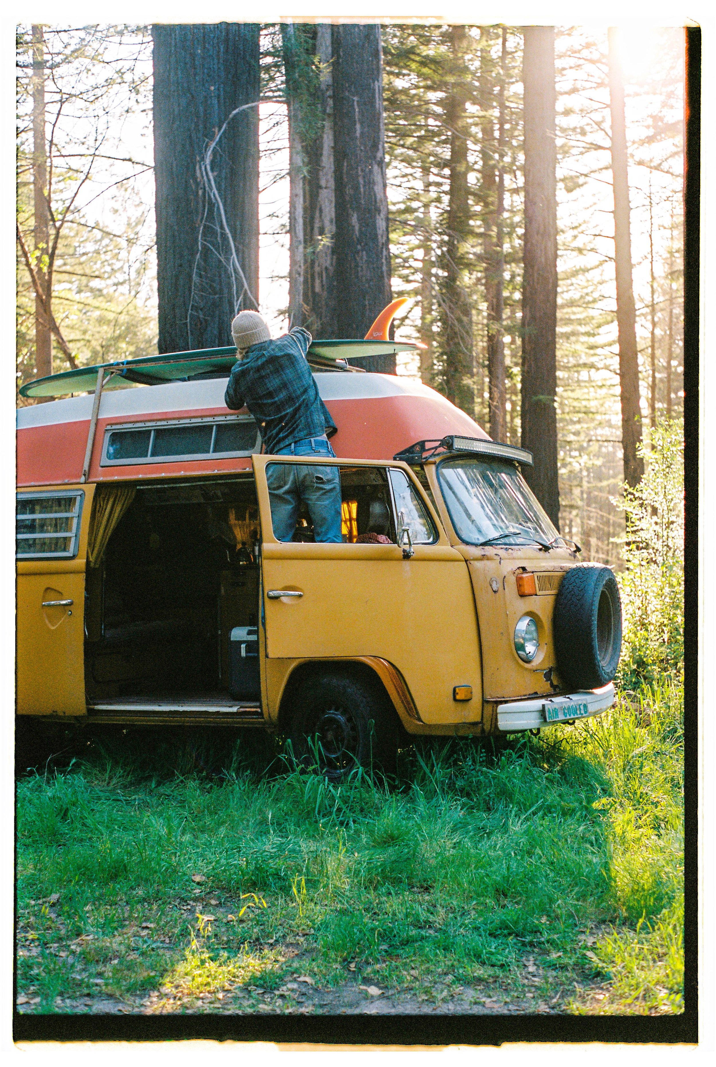 A person placing a surfboard on top of a vintage yellow campervan in a forest with tall trees during sunlight.
