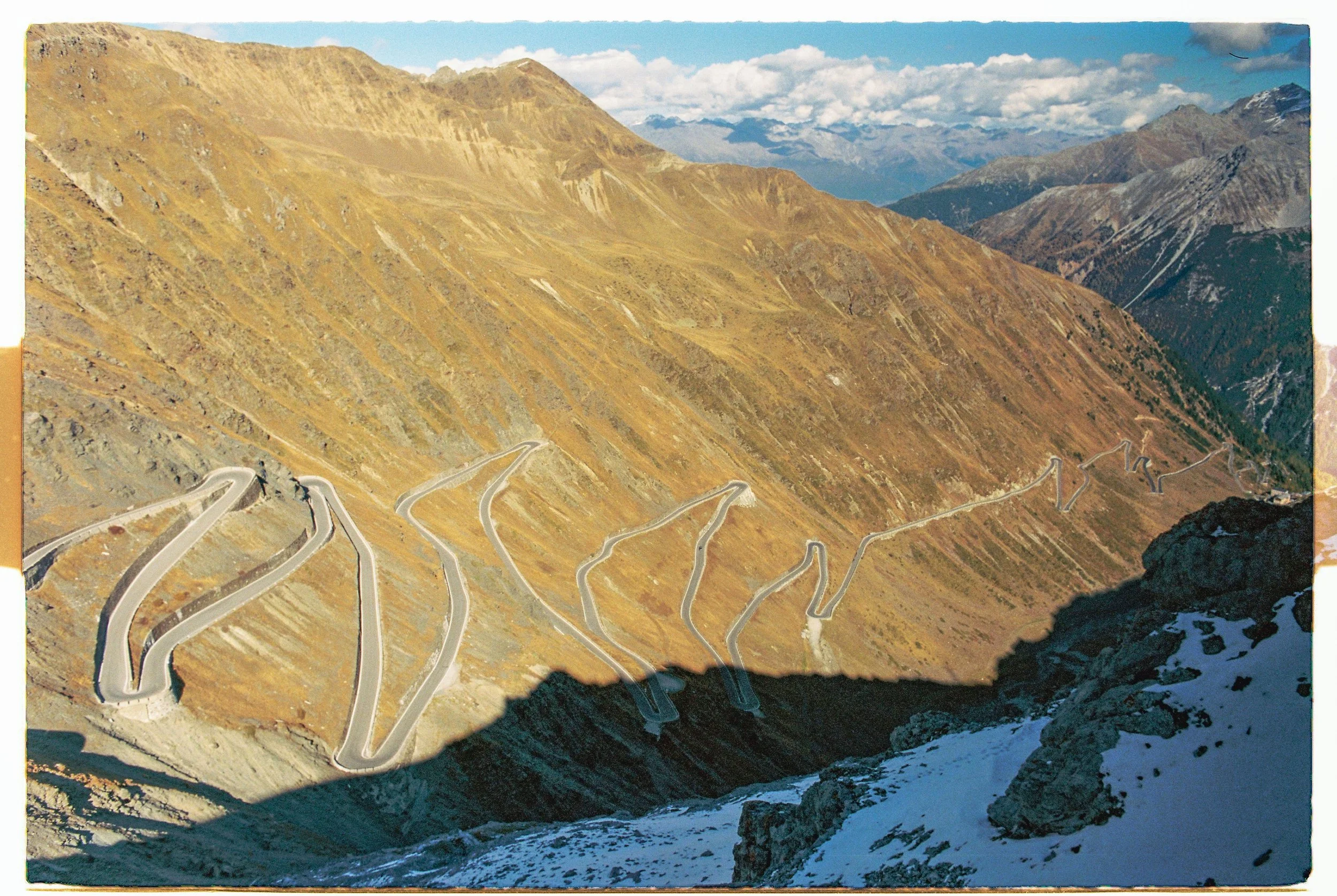 A winding mountain road with multiple switchbacks, seen from a high vantage point overlooking a valley with distant mountains, some snow on the ground and a cloudy sky.