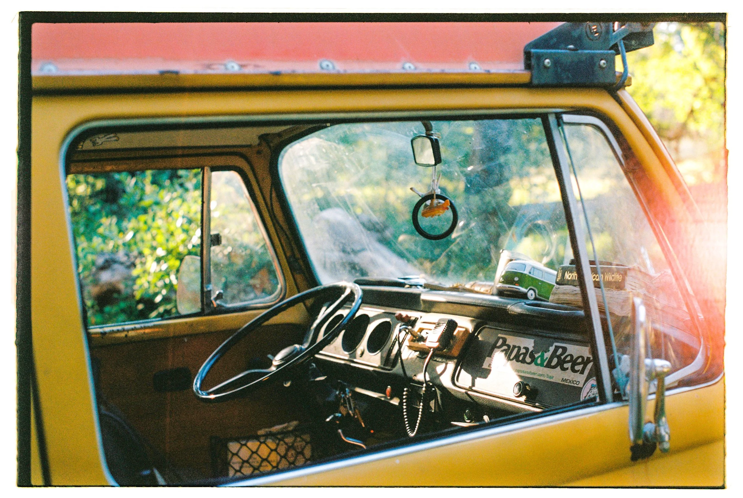 Inside view of a vintage yellow vehicle's dashboard and windshield with various souvenirs and stickers, including a 'Road & Beer' sticker, a miniature Volkswagen bus, and a hanging air freshener, with green foliage outside.