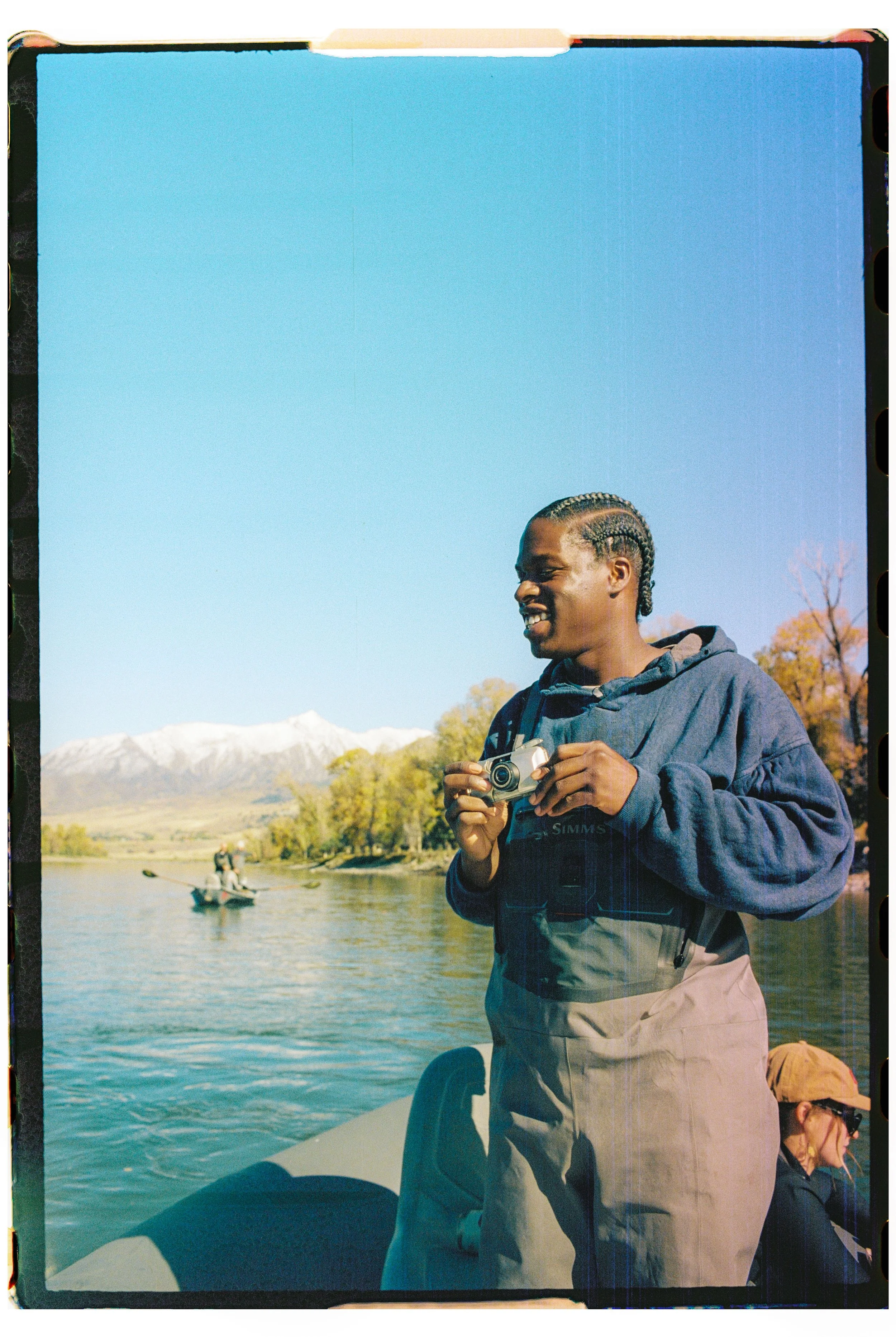 Person holding a camera on a boat with a lake and mountains in the background, and trees with autumn leaves.