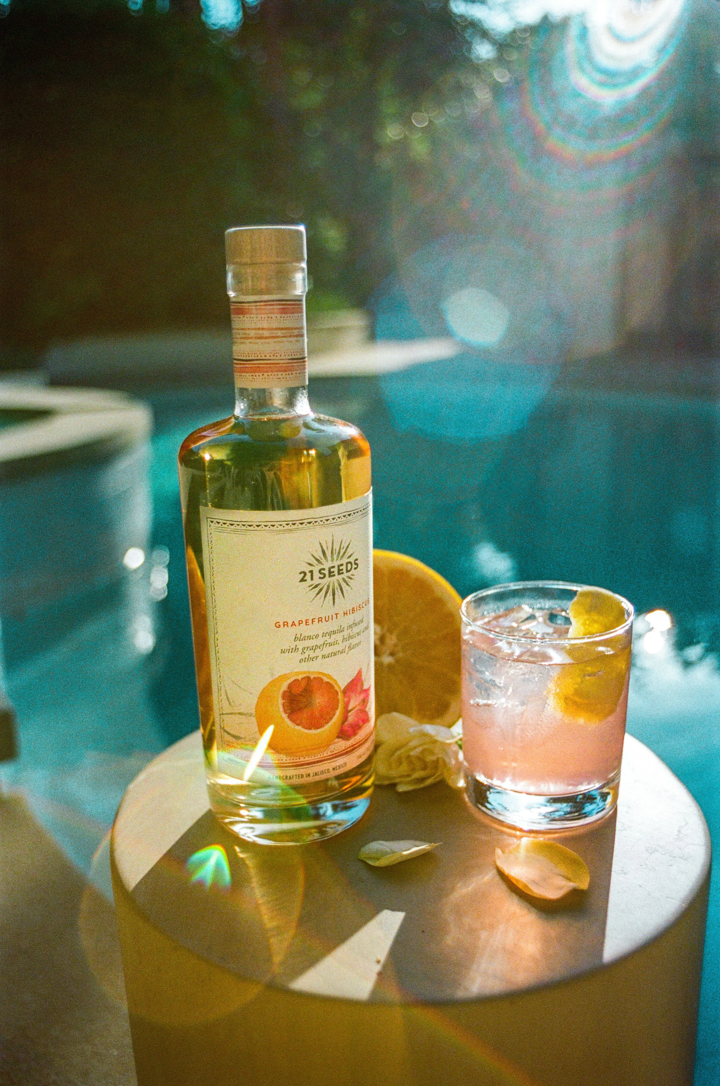 A bottle of 21 Seeds grapefruit hibiscus tequila and a pink cocktail with lemon slices on a small round table by a pool, surrounded by poolside decor with sunlight and rainbow lens flare.