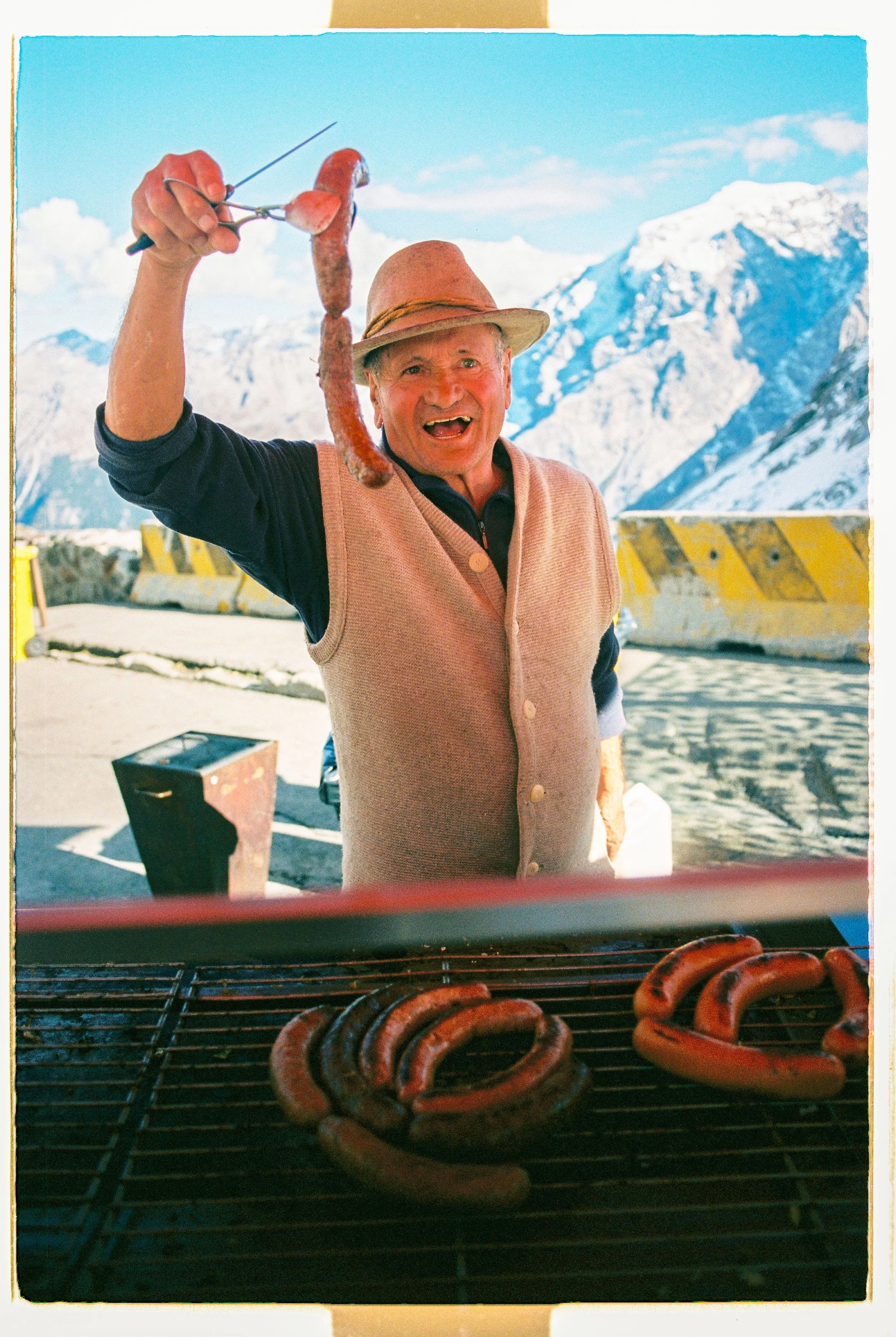 A man wearing a hat and vest grills sausages outdoors with snowy mountains in the background. He is holding a sausage with a skewer, smiling, and standing behind a grill with more sausages.