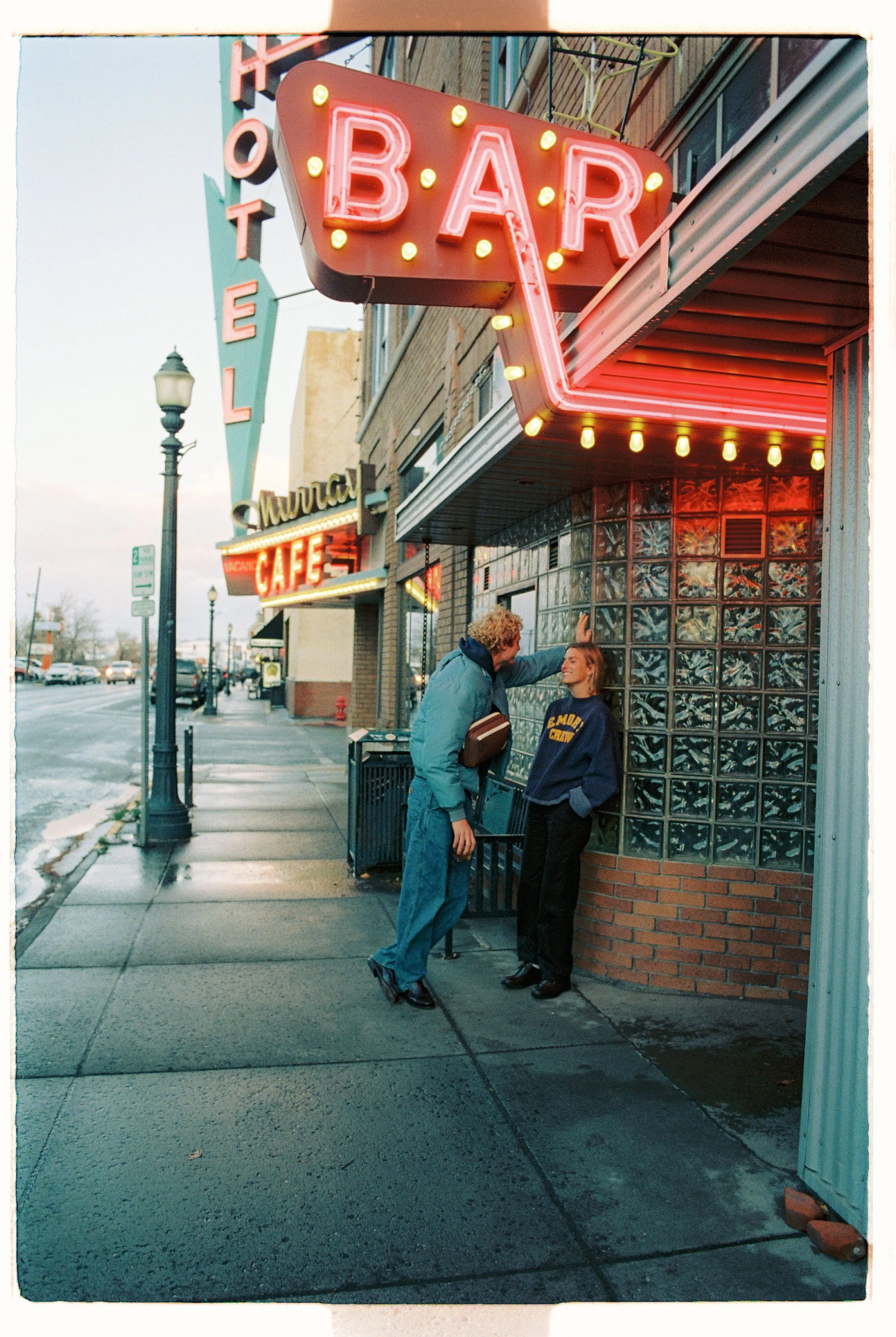 A man and woman standing outside a neon-lit bar and cafe on a city sidewalk, with a vintage atmosphere and streetlights.