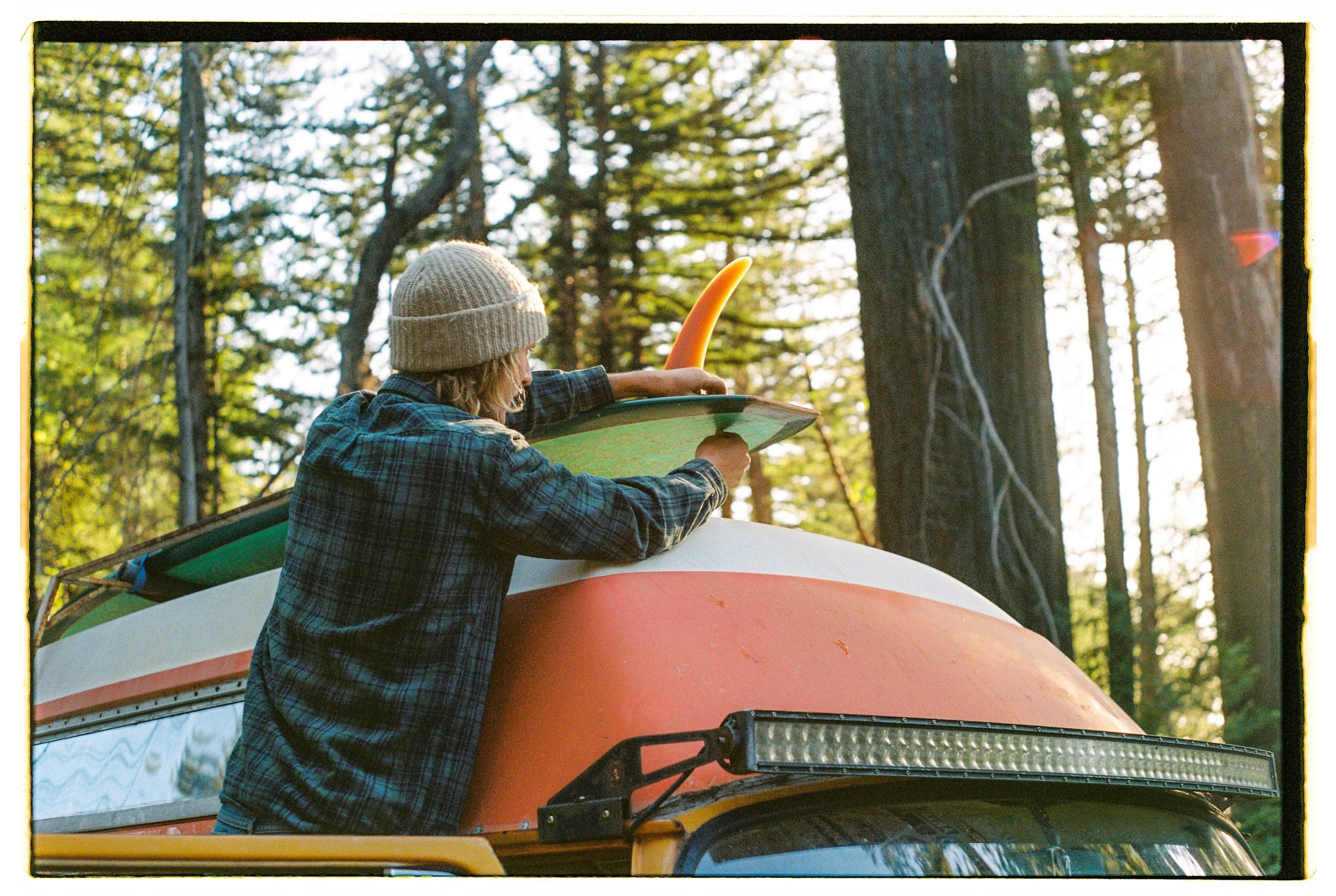 A person loading a surfboard onto a vehicle in a forested area during the daytime.