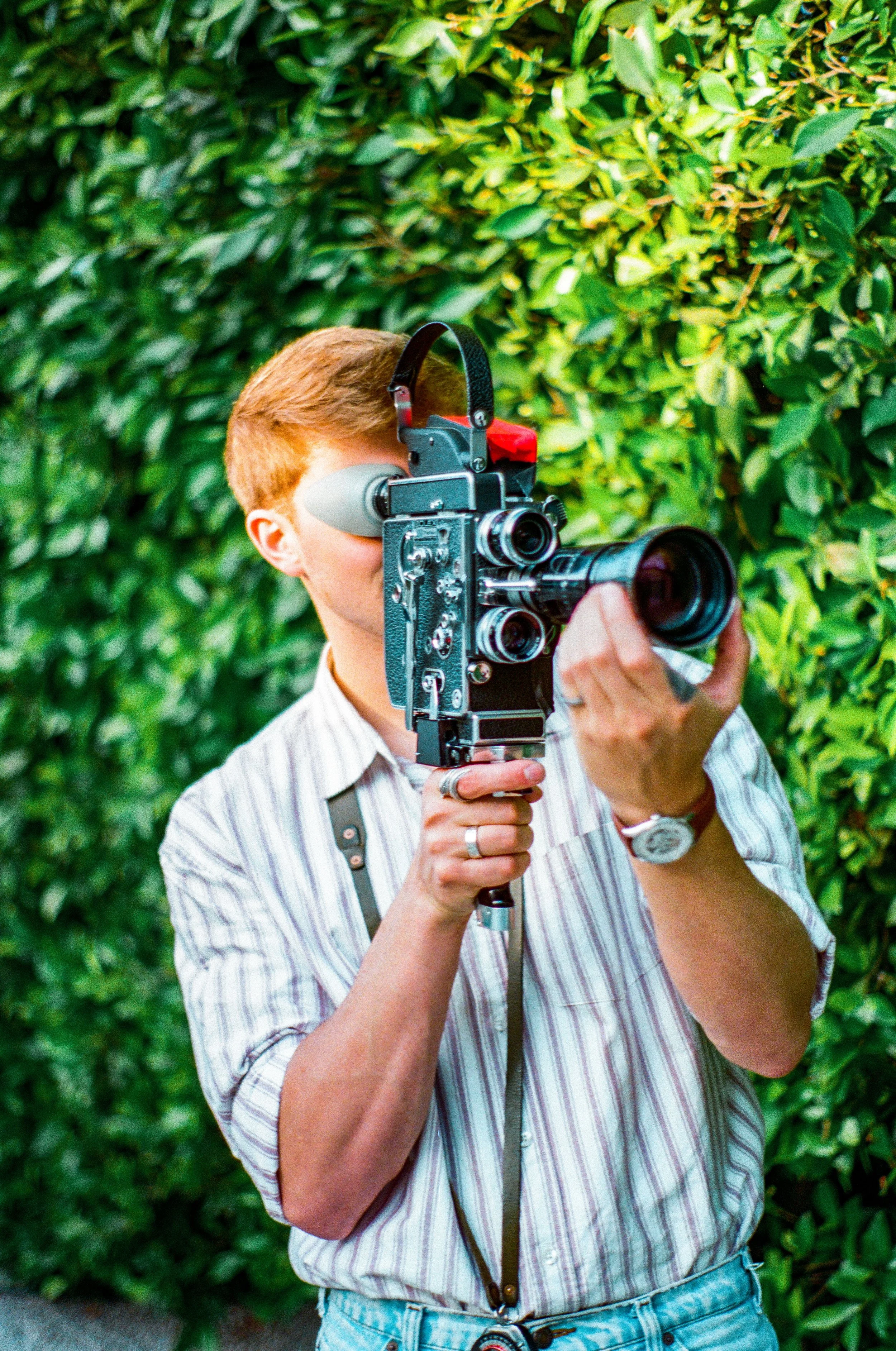A man with red hair and a striped shirt aiming a professional video camera at a green leafy background.
