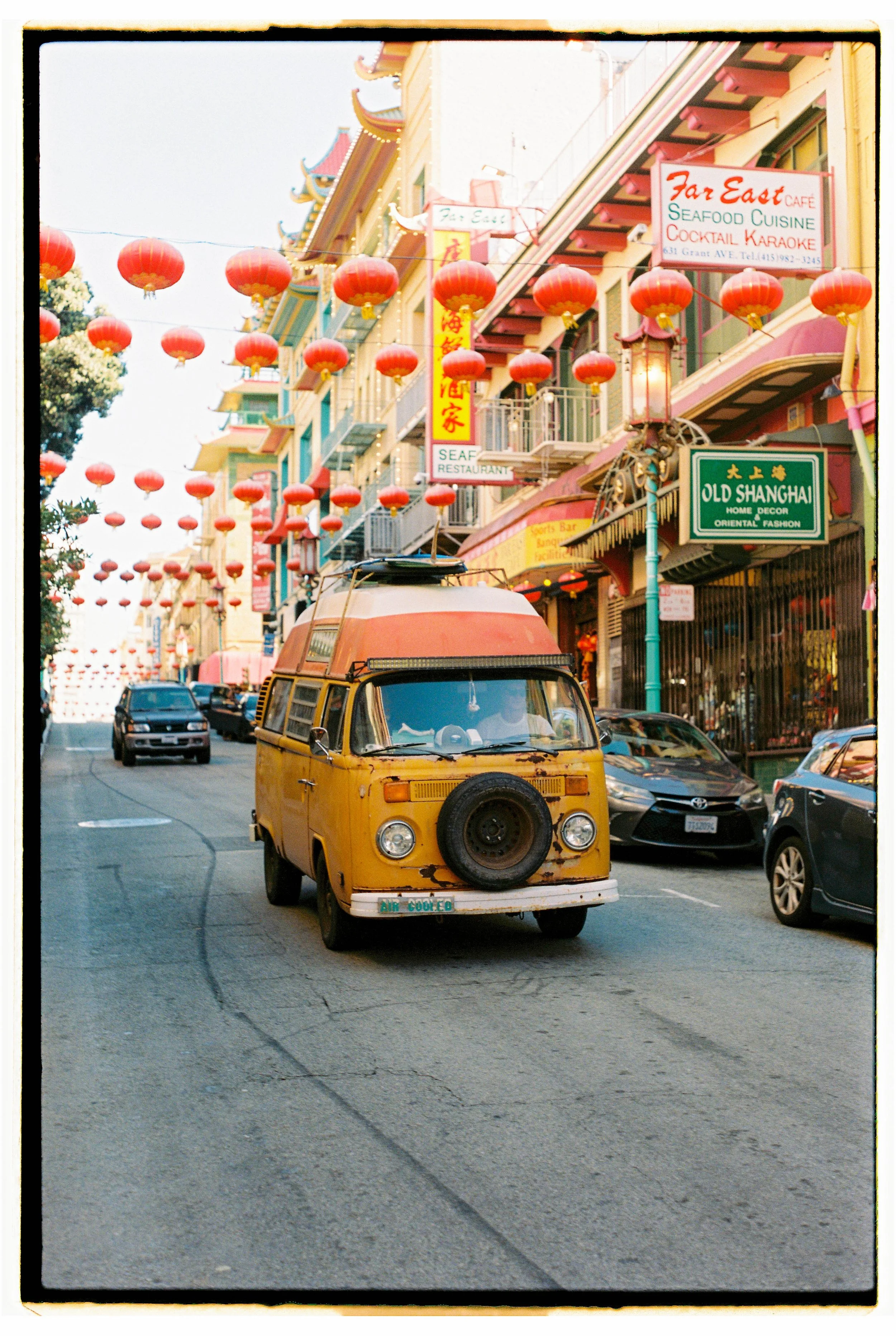A yellow vintage Volkswagen camper van with a spare tire on the front, driving down a street decorated with red Chinese lanterns. The buildings are colorful with signs for Asian restaurants and shops, including 