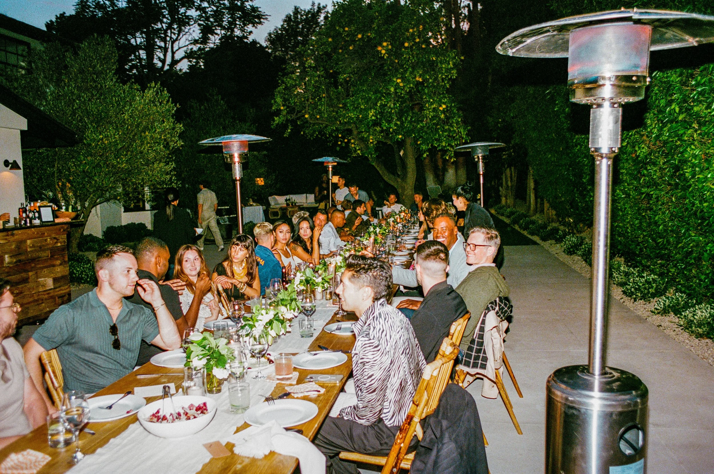 A large outdoor dinner party with many people seated at a long table set with white tablecloths, plates, glasses, and greenery centerpieces, under multiple patio heaters, in a garden setting during evening.