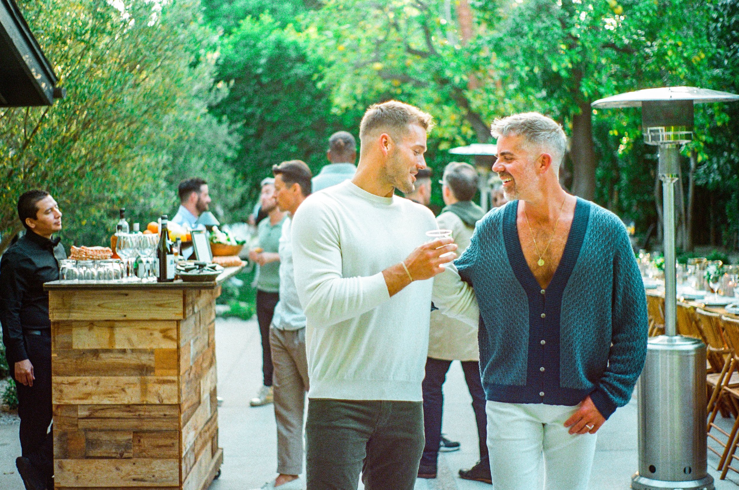 Two men smiling and talking at an outdoor gathering during daytime, with a wooden bar and other guests in the background
