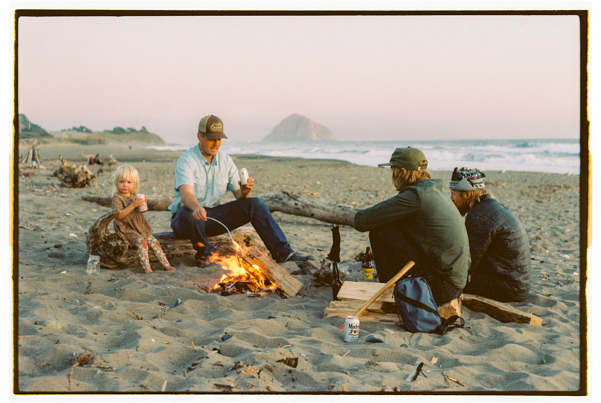 Four people sitting around a small campfire on a beach at sunset, with a young girl, two men, and a woman, surrounded by driftwood and drinks, with ocean waves and a rock formation in the background.