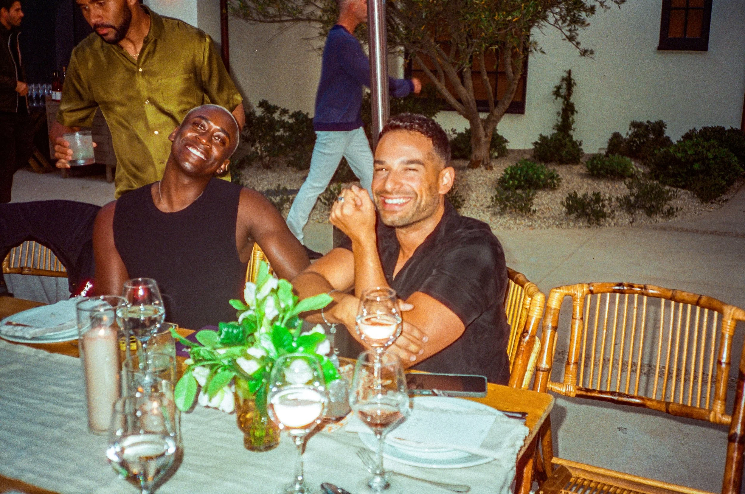 Two men smiling and enjoying themselves at a dinner table outdoors, with table settings and a floral centerpiece. Others are visible in the background at a social gathering.