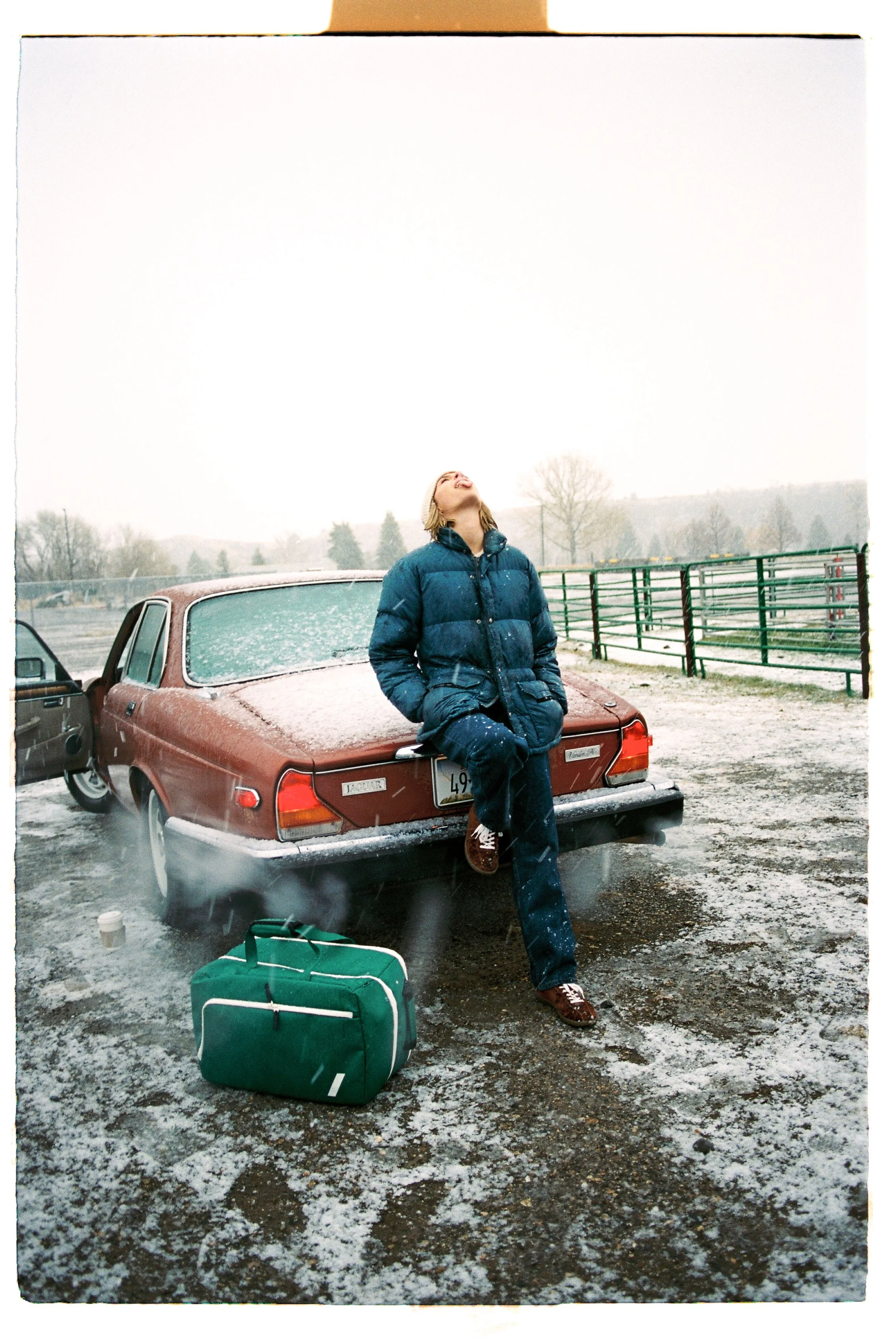 Person in a blue jacket leaning against the back of a dusty red classic car, sitting on the trunk, with an open door nearby, in an outdoor snowy parking lot.