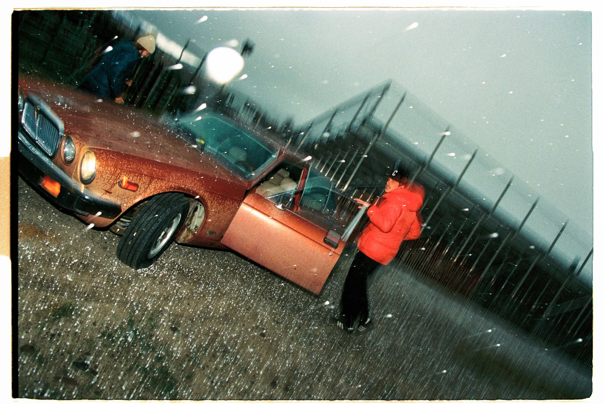 Person in red jacket standing next to a vintage brown car on a rainy day with a fence and utility poles in the background.