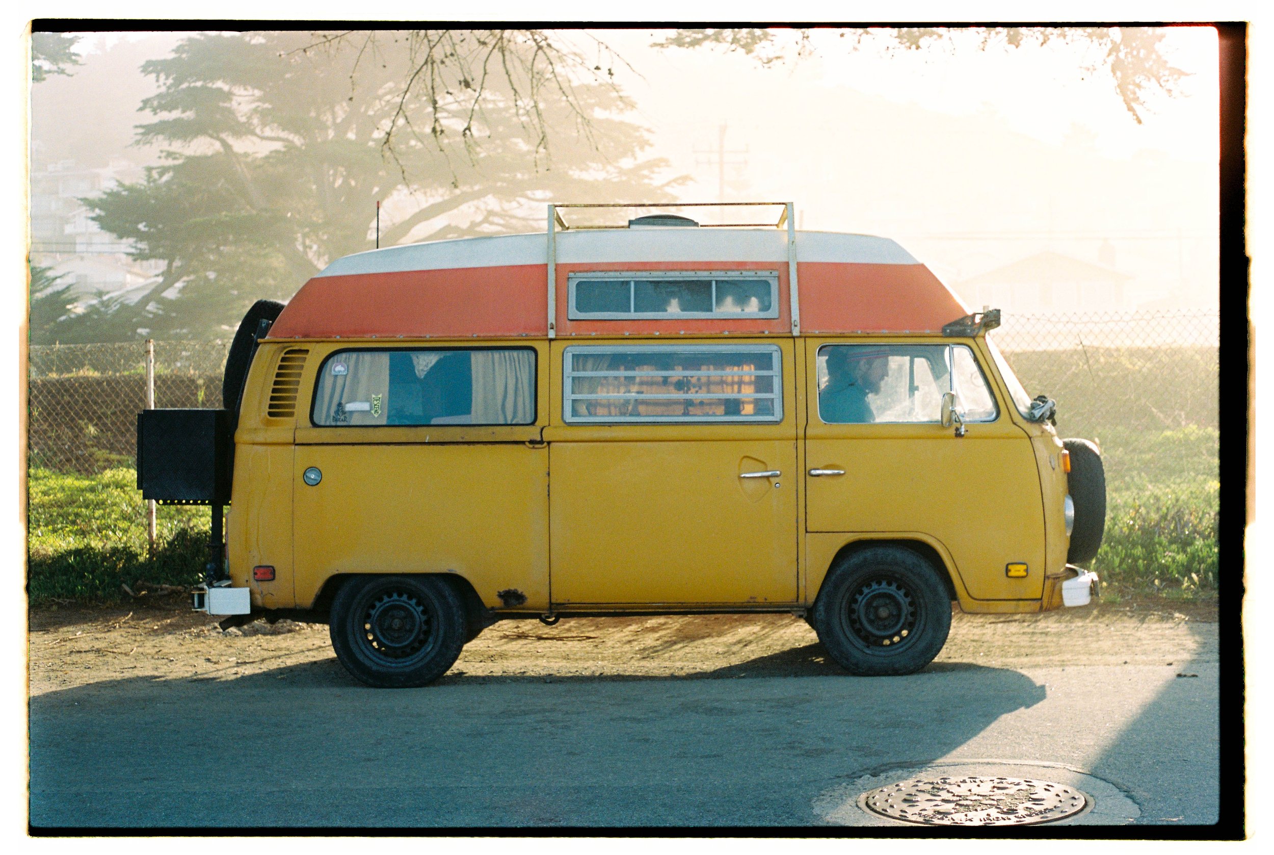 A vintage yellow Volkswagen camper van with a red and white roof parked on the side of a street, with a chain-link fence and trees in the background, and the sun casting shadows.