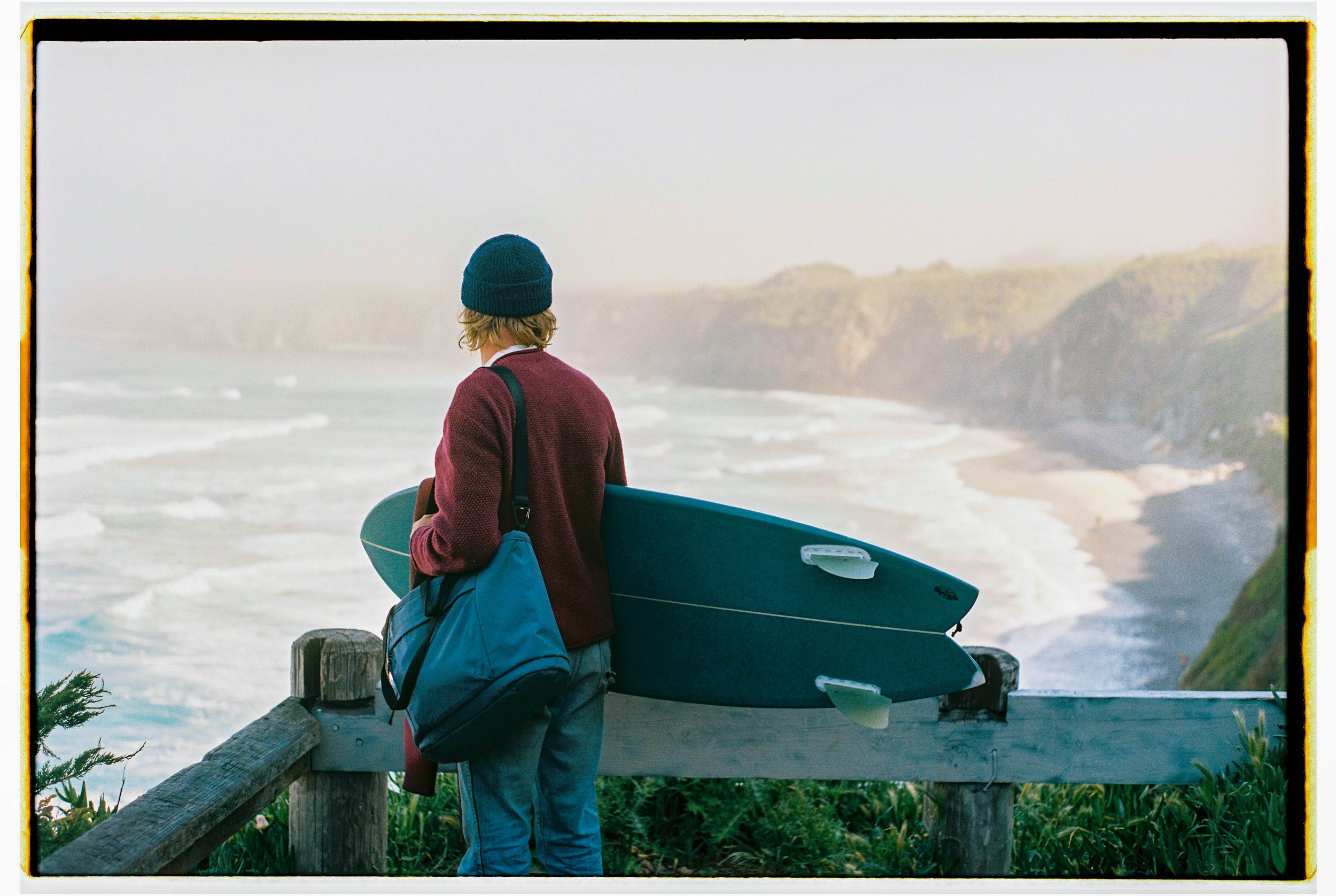 A person with blonde hair wearing a blue beanie, maroon sweater, and carrying a black and blue bag stands on a wooden viewing platform, holding a surfboard, looking out at a foggy coastal landscape with waves and cliffs.