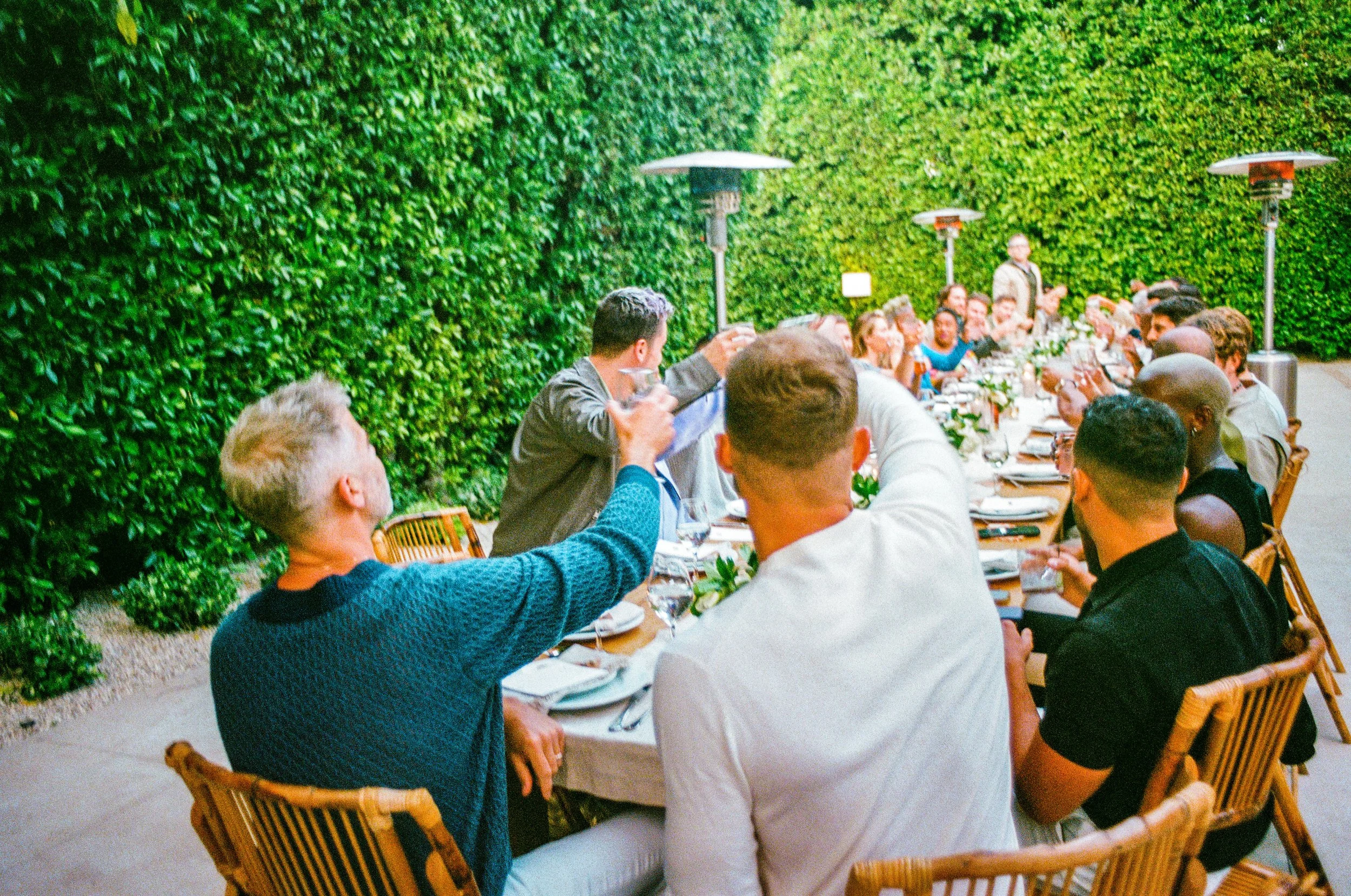 People gathered at a long outdoor dining table surrounded by green hedges, raising glasses in a toast.