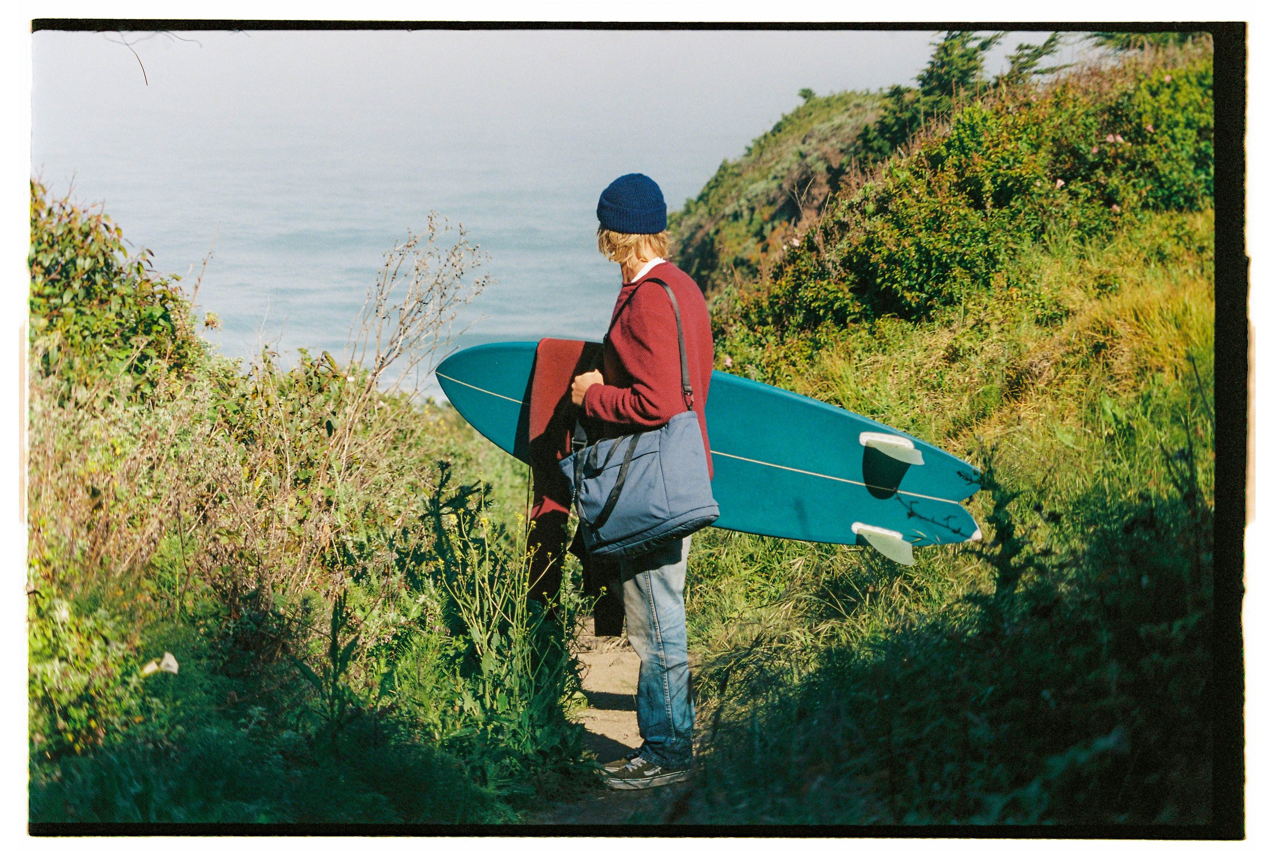 Person dressed in a maroon jacket and blue beanie holding a surfboard, standing on a trail near the coast with green shrubbery and ocean in the background.
