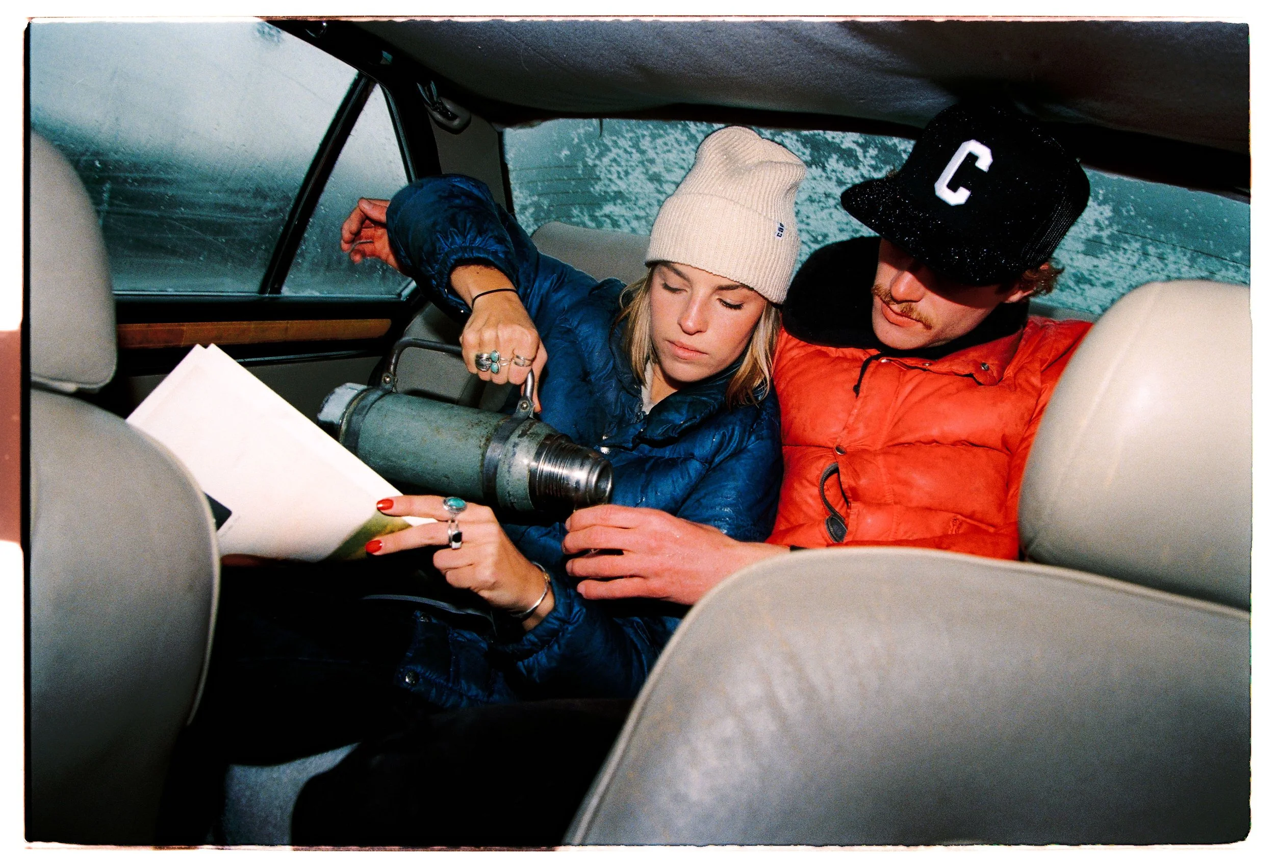 A woman and a man sitting in the backseat of a car during a rainy day, with the woman holding a thermos and a paper, both dressed in casual jackets and beanies.