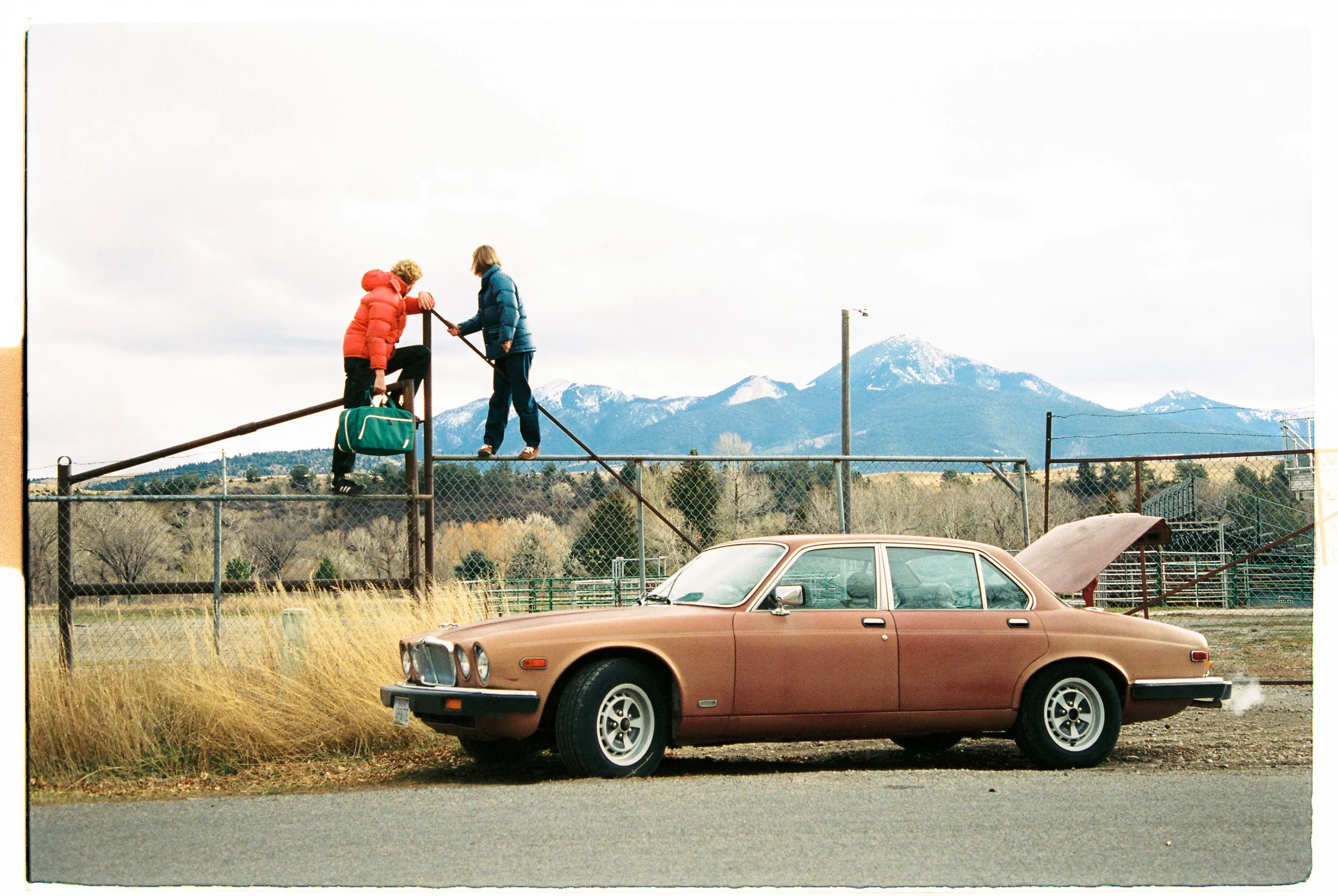 Two children on a playground with a mountain range in the background, one climbing a metal structure and the other handing something to them; a vintage brown car parked nearby.