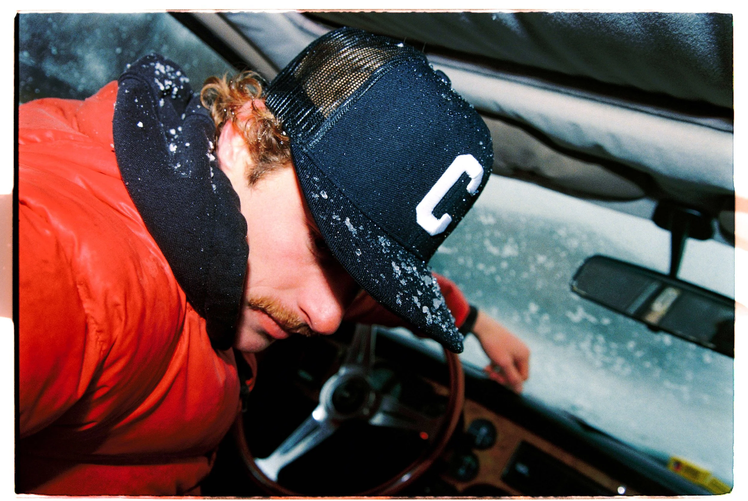 A person with reddish hair and a mustache wearing a black cap with a white letter 'C', a black hoodie, and an orange puffer jacket, sleeping or resting in the driver's seat of a vehicle during a snowstorm.