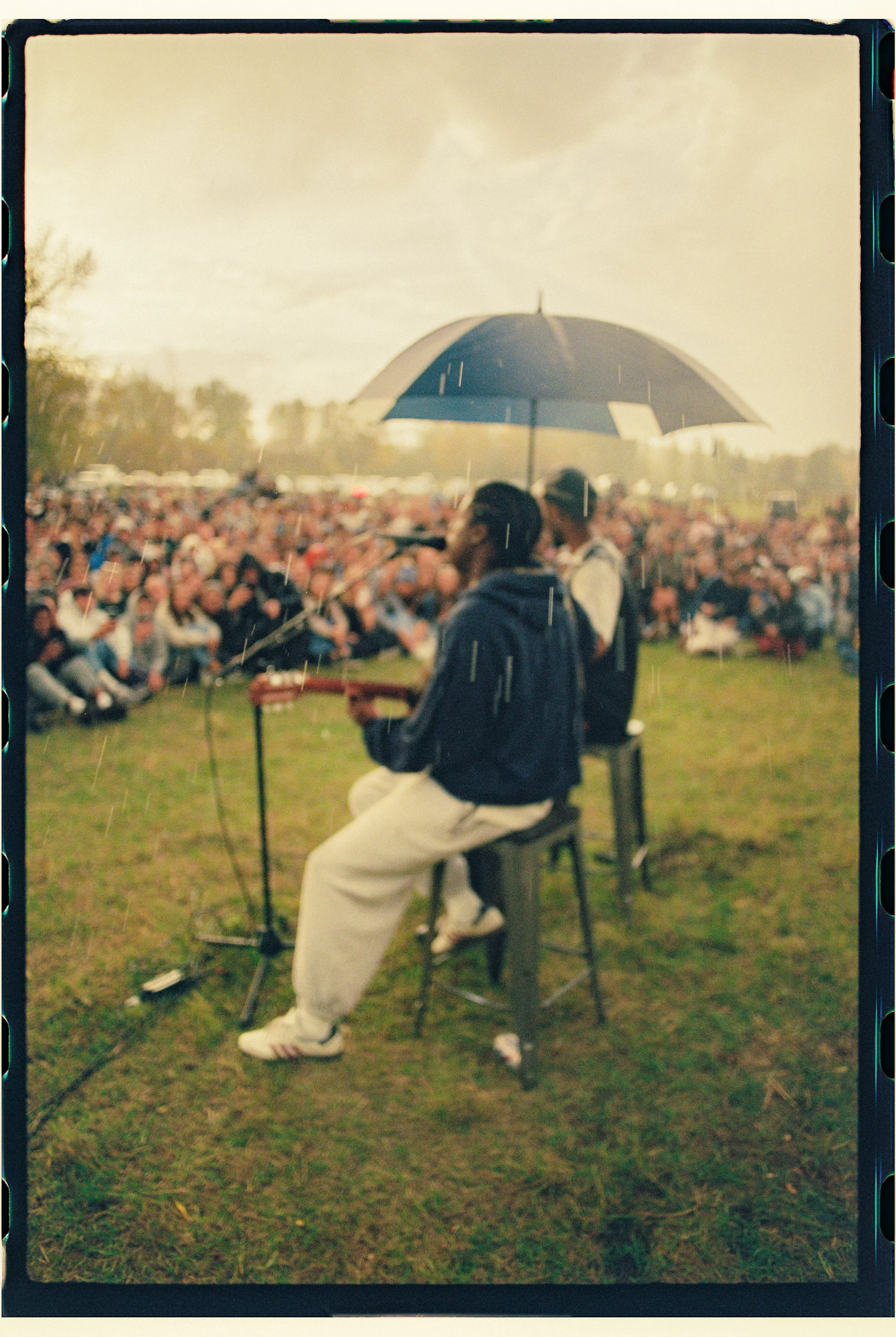 Musicians performing with one holding an umbrella in front of a large outdoor audience during rain.