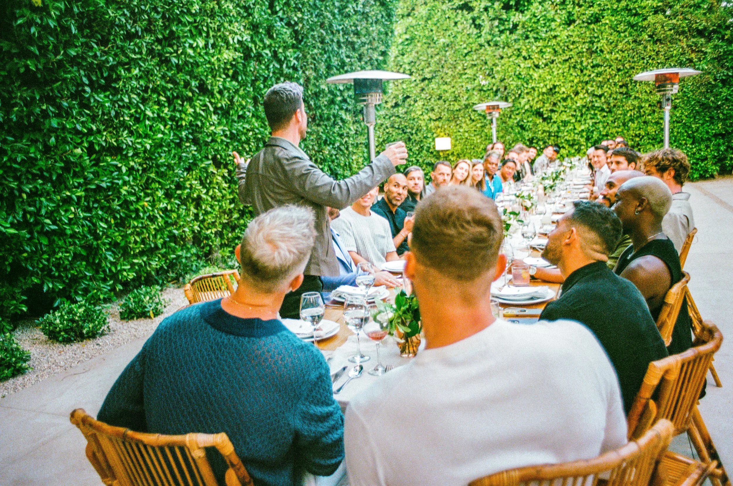 People gathered at an outdoor dinner party, listening to a man giving a toast or speech, with a green hedge background and heaters overhead.