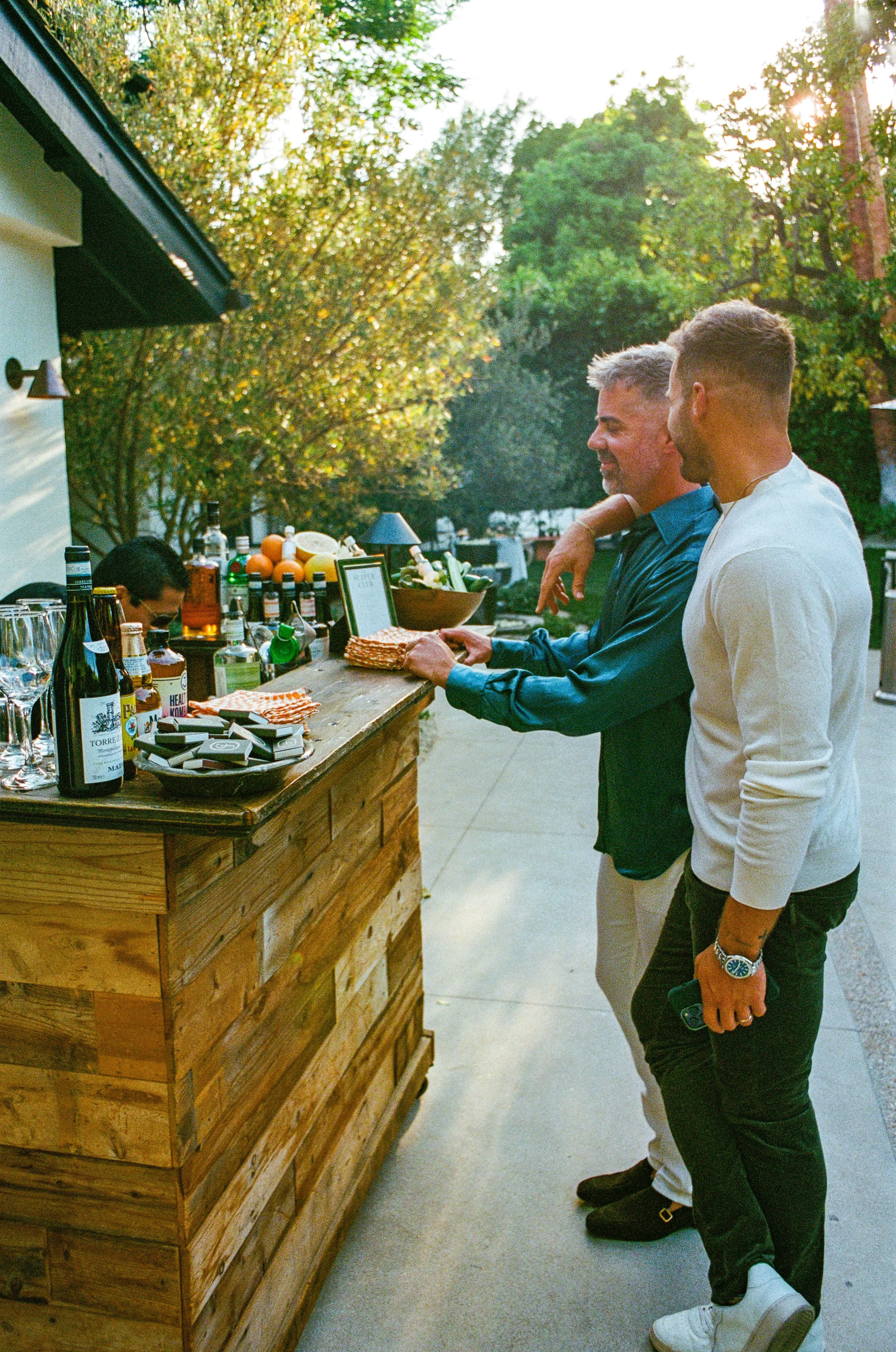 Two men stand at an outdoor bar with a variety of drinks and snacks, engaging in conversation, surrounded by trees and warm sunlight.