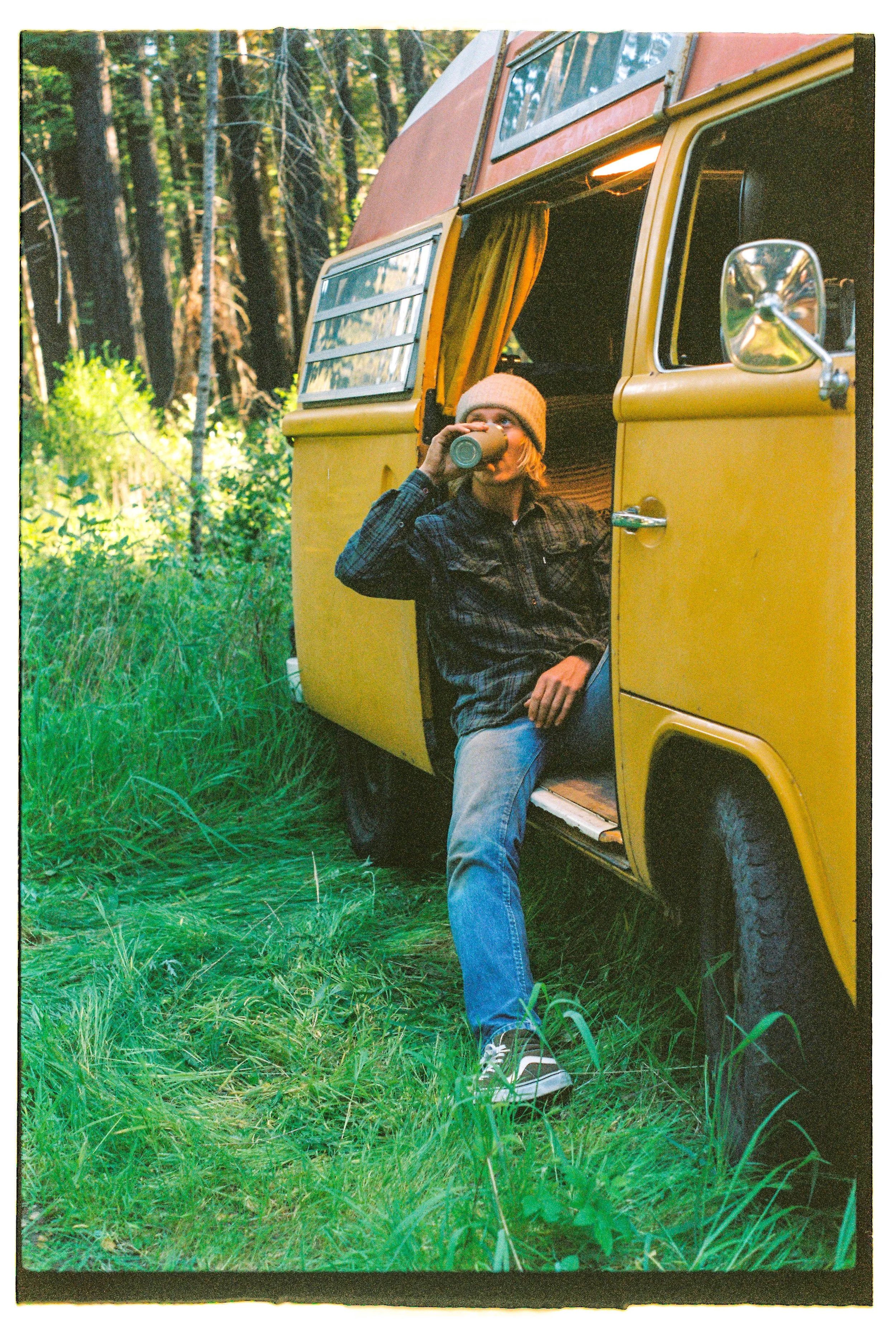 A person sitting at the open door of a yellow camper van, drinking from a cup in a forest setting.