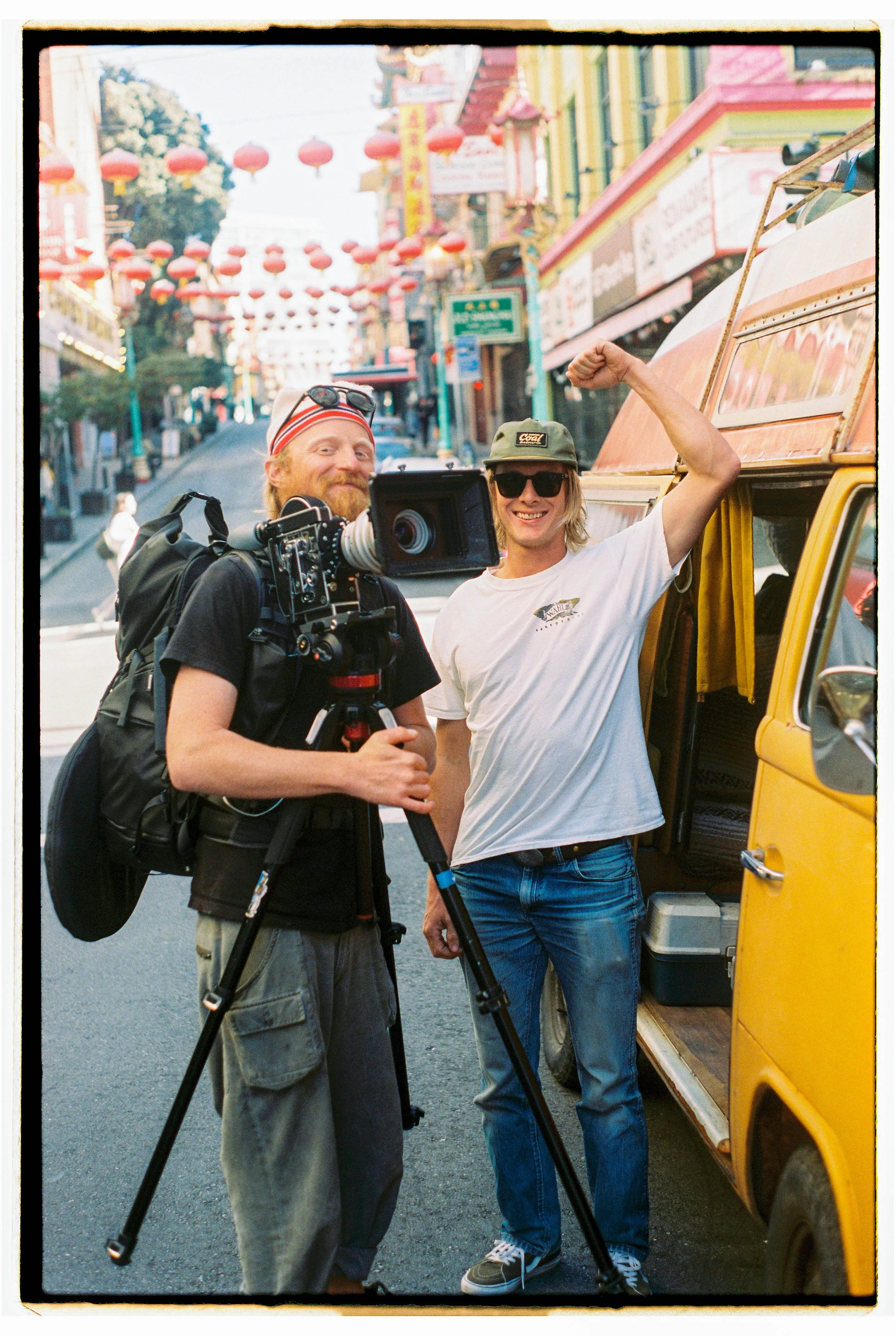 Two men stand on a street with colorful buildings and lanterns. One man holds a video camera, and the other is flexing his arm muscle while smiling.