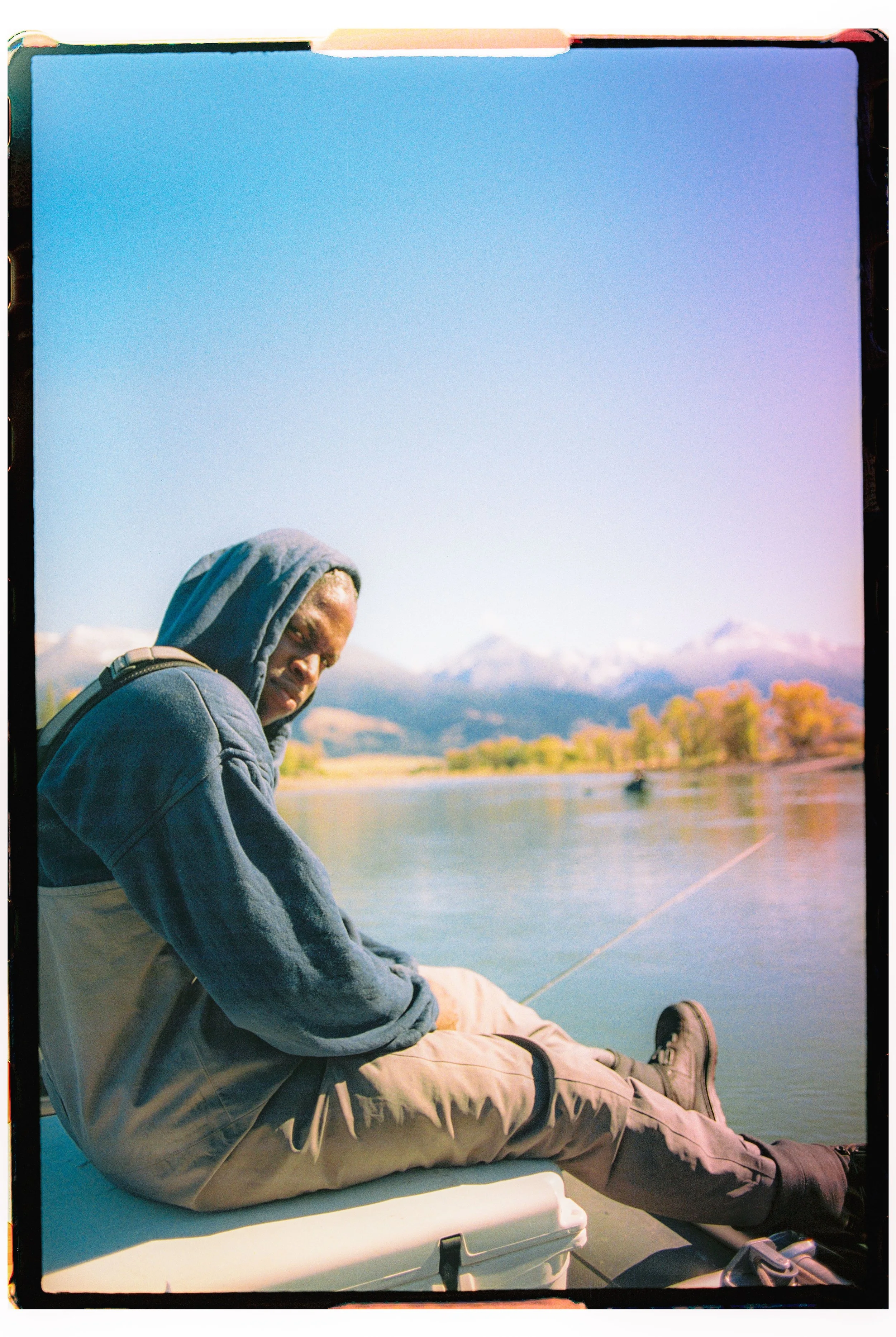 A man in a hoodie and outdoor clothing sitting on a boat by a river, fishing with mountains and trees in the background.