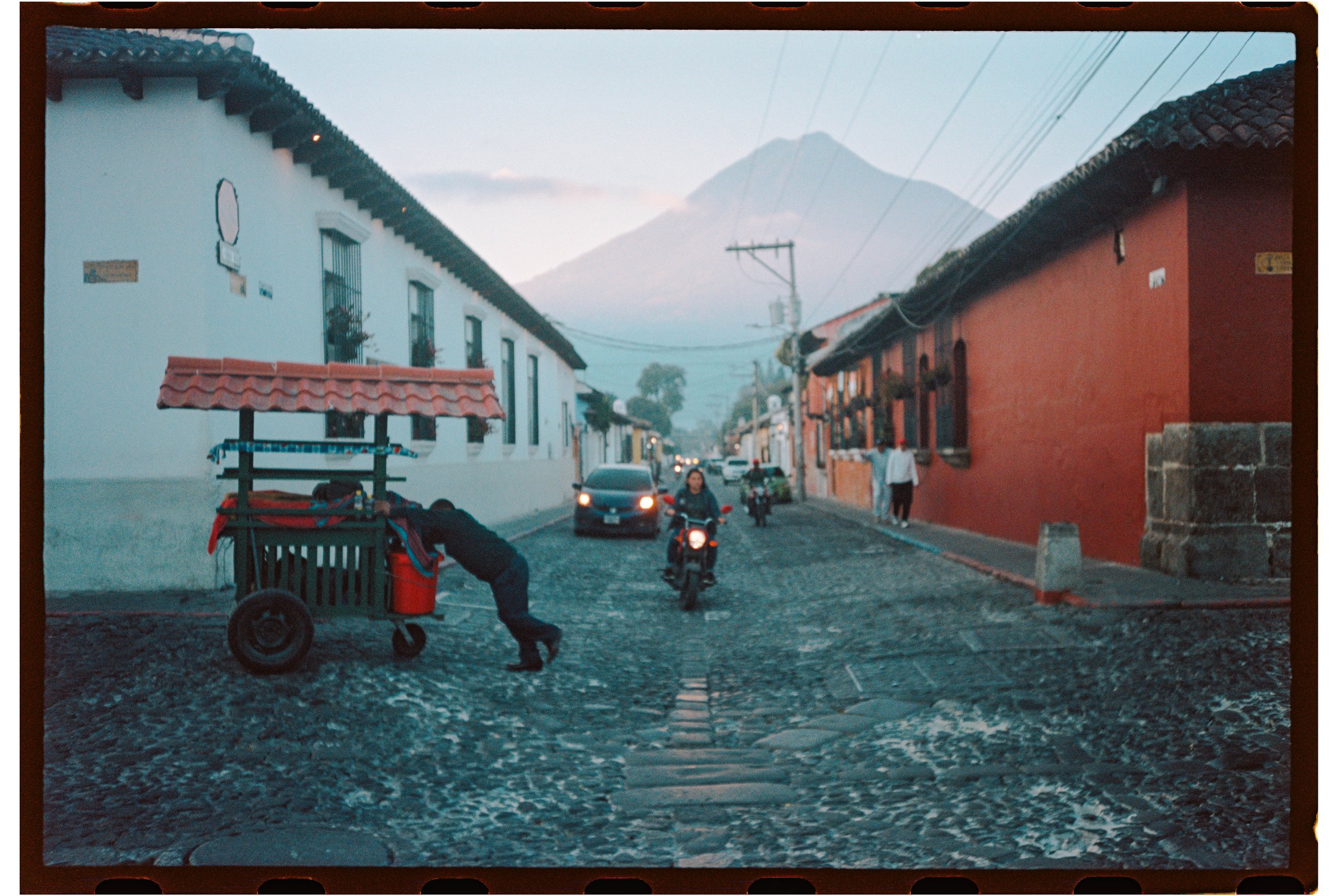 Cobblestone street in a town with colorful colonial-style buildings, a volcano in the background, motorcycles and cars on the street, and a street vendor cart in the foreground.