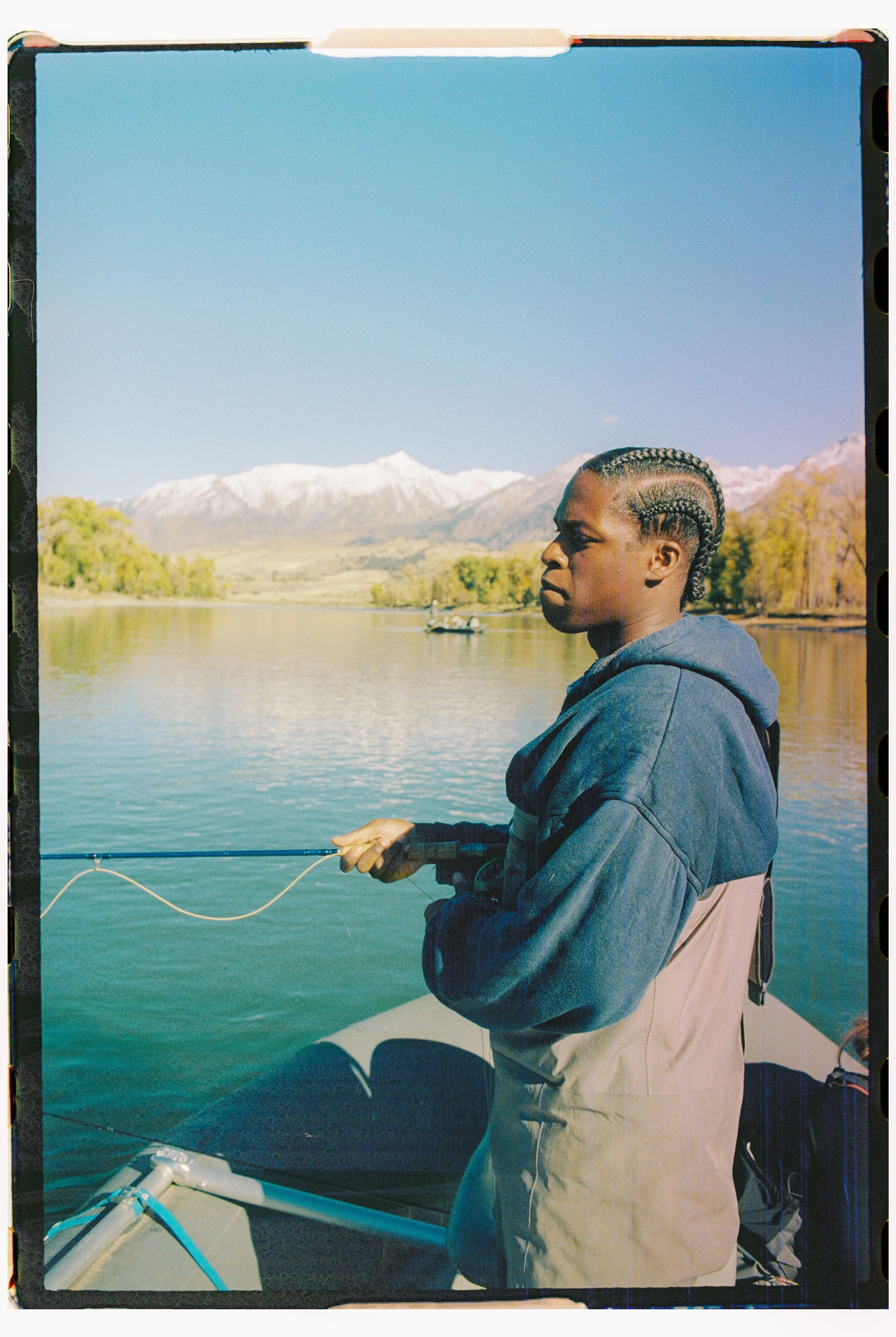 A person standing in a boat on a lake, fishing with a mountain range in the background, and snow-capped peaks under a clear blue sky.