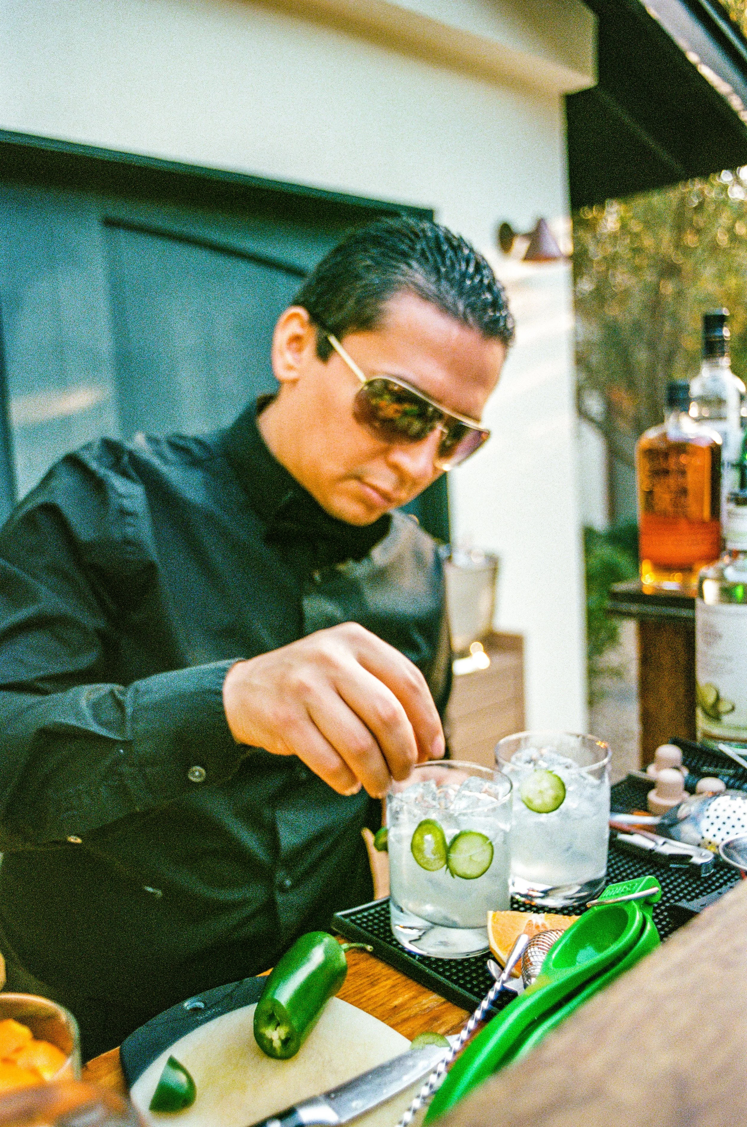 A man wearing sunglasses and a black shirt preparing cocktails with slices of cucumber in clear glasses on a bar counter.