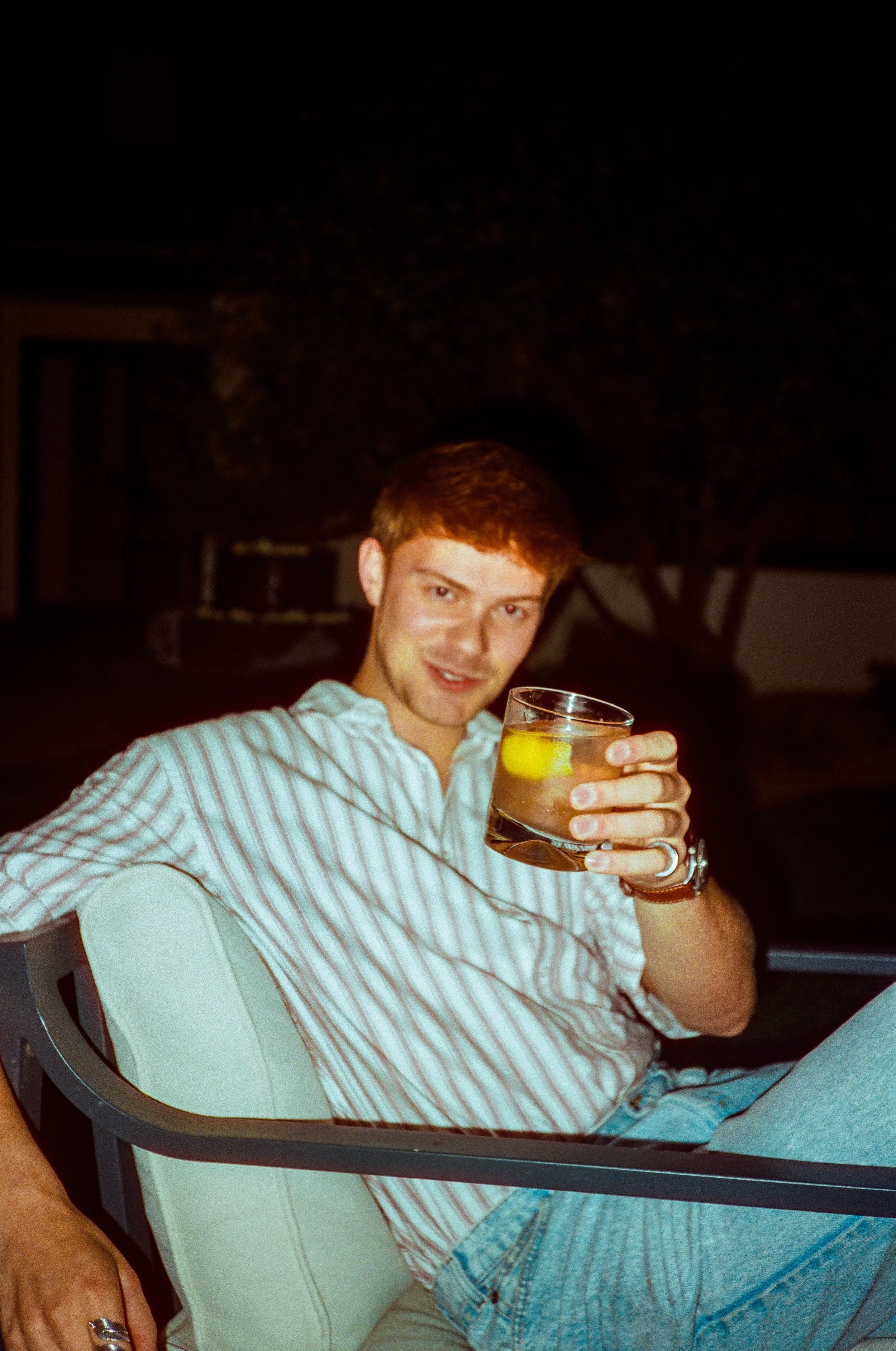 A young man sitting outdoors at night, holding a glass of beverage with lemon slices, and smiling at the camera.