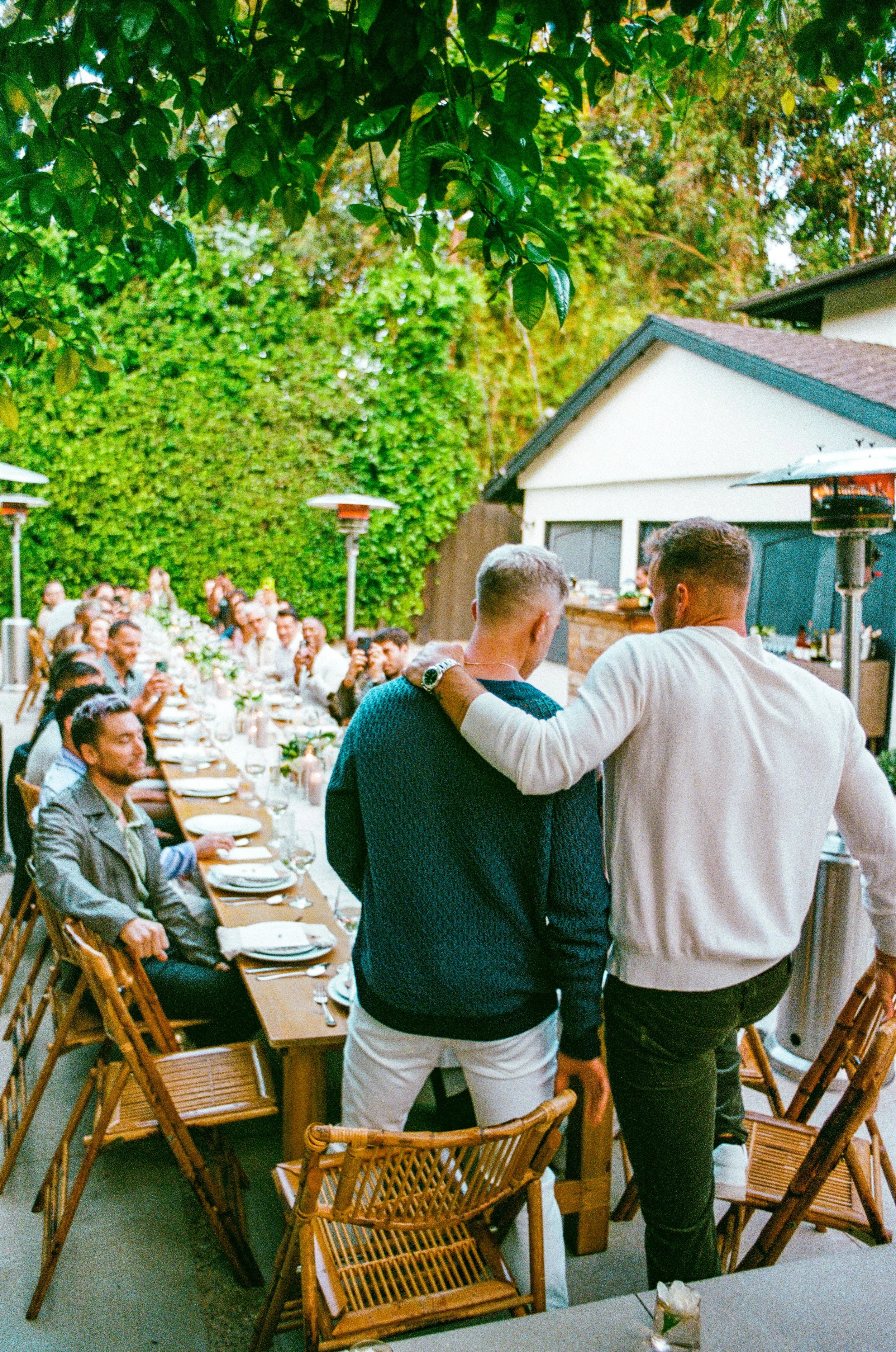 People gathered around a long outdoor dining table, with two men standing and talking in the foreground, at a garden party or outdoor celebration.