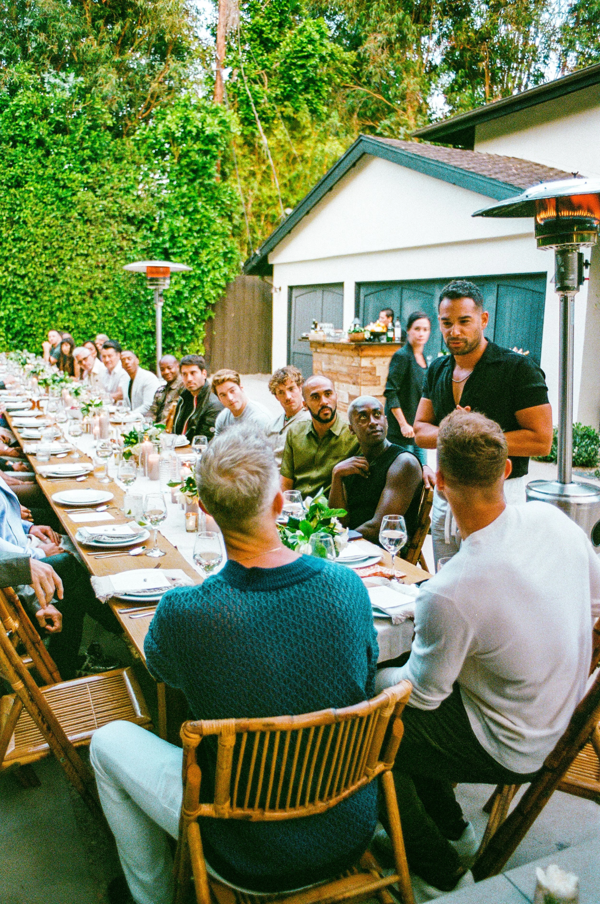 A large outdoor dinner gathering with people sitting at a long table with white tablecloths, plates, glasses, and floral centerpieces, in a lush backyard with trees and a garage, and a man standing and speaking to the group.