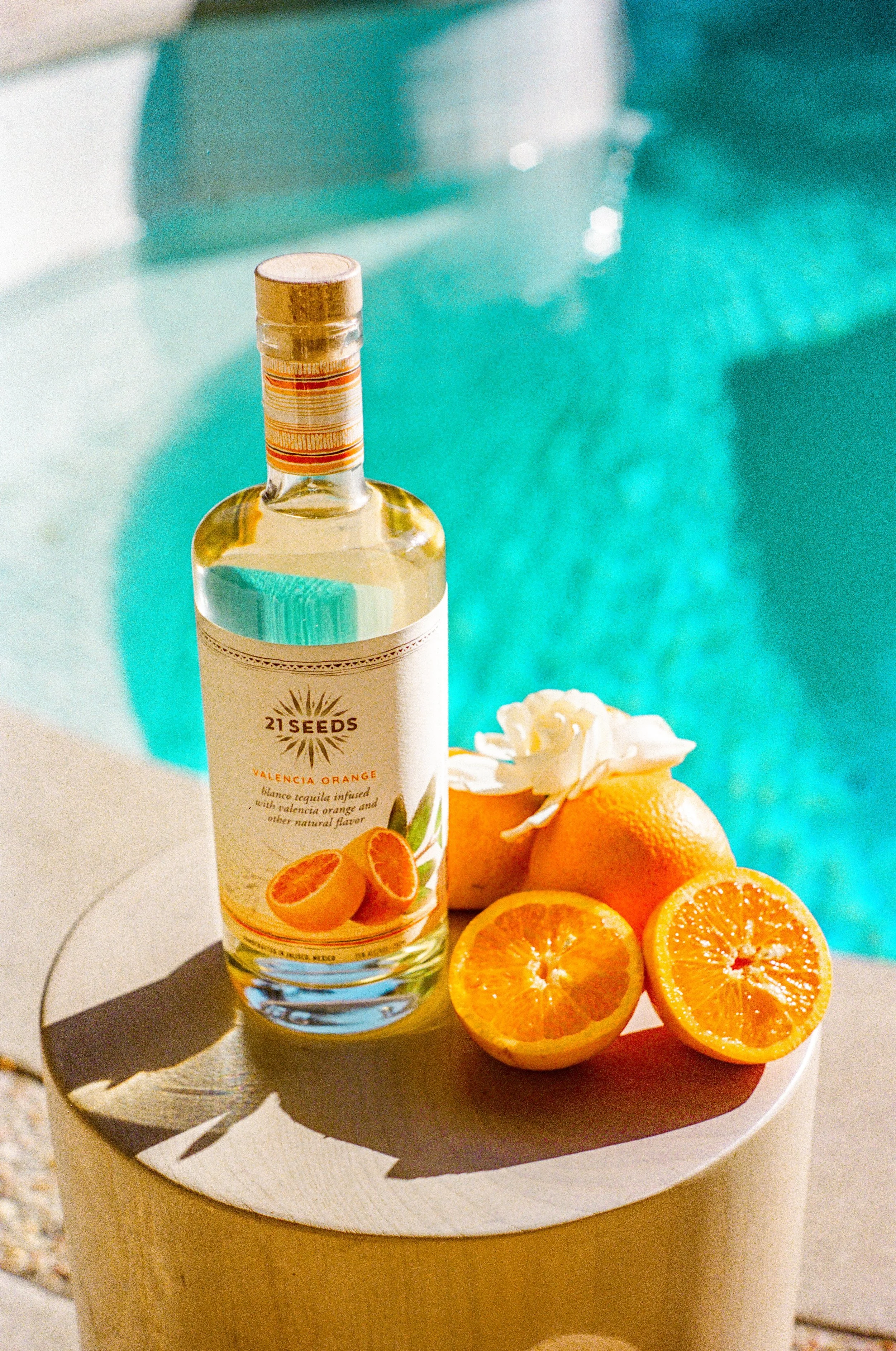 A bottle of 21 Seeds Valencia Orange tequila infused with natural orange flavor, placed on a white circular table next to fresh cut oranges and a white flower, with a pool in the background.